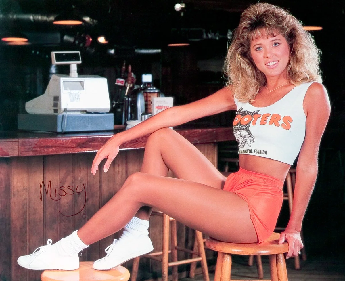 A Hooters Calendar Girl with curly blonde hair sitting on a wooden barstool in a Hooters uniform, consisting of a white crop top with the Hooters logo and orange shorts, smiling at the camera inside a bar or restaurant.