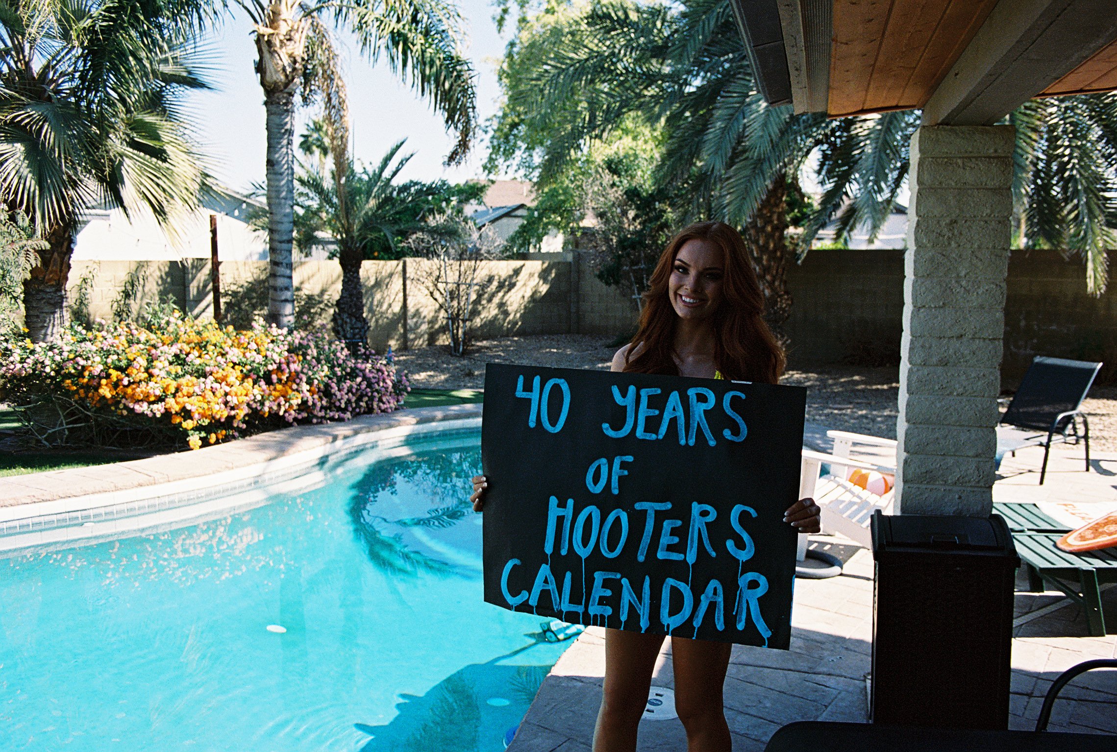 Woman with red hair holding a black sign that reads '40 Years of Hooters Calendar' by a backyard pool with palm trees and colorful flowers.