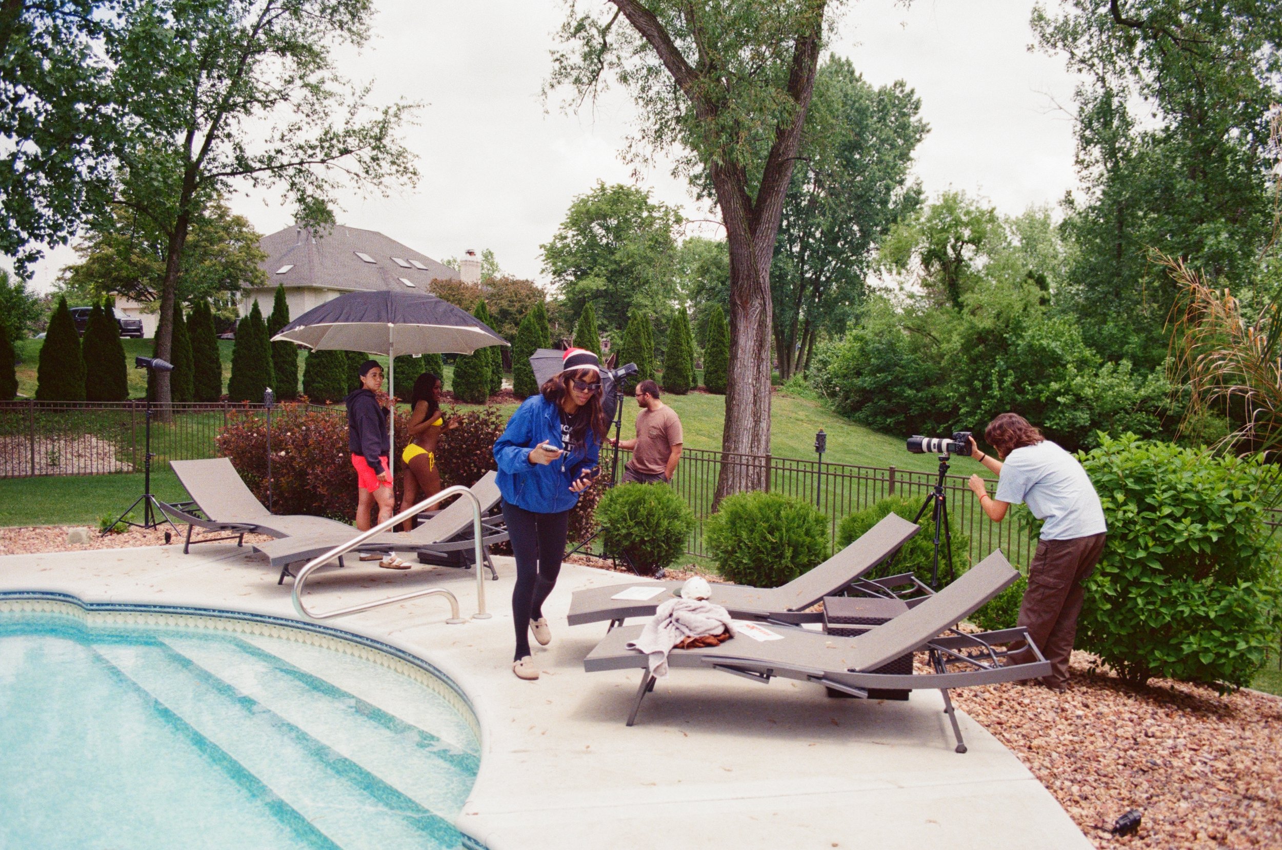Group of people preparing for a photo shoot by a swimming pool with lounge chairs, umbrellas, and lush greenery in the background.