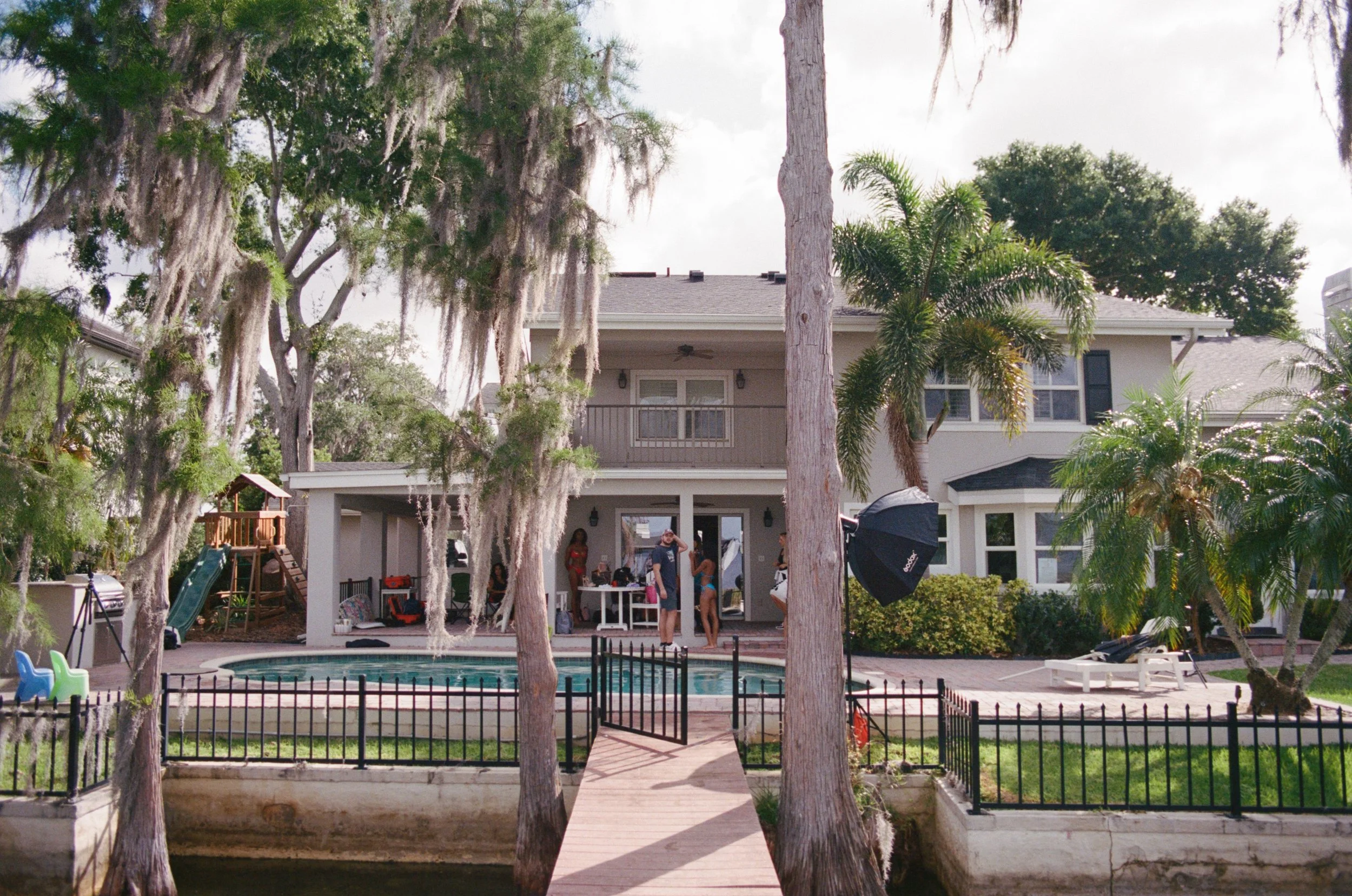 A backyard view of a two-story house with a pool, surrounded by trees and greenery. People are gathered on the patio near the pool.