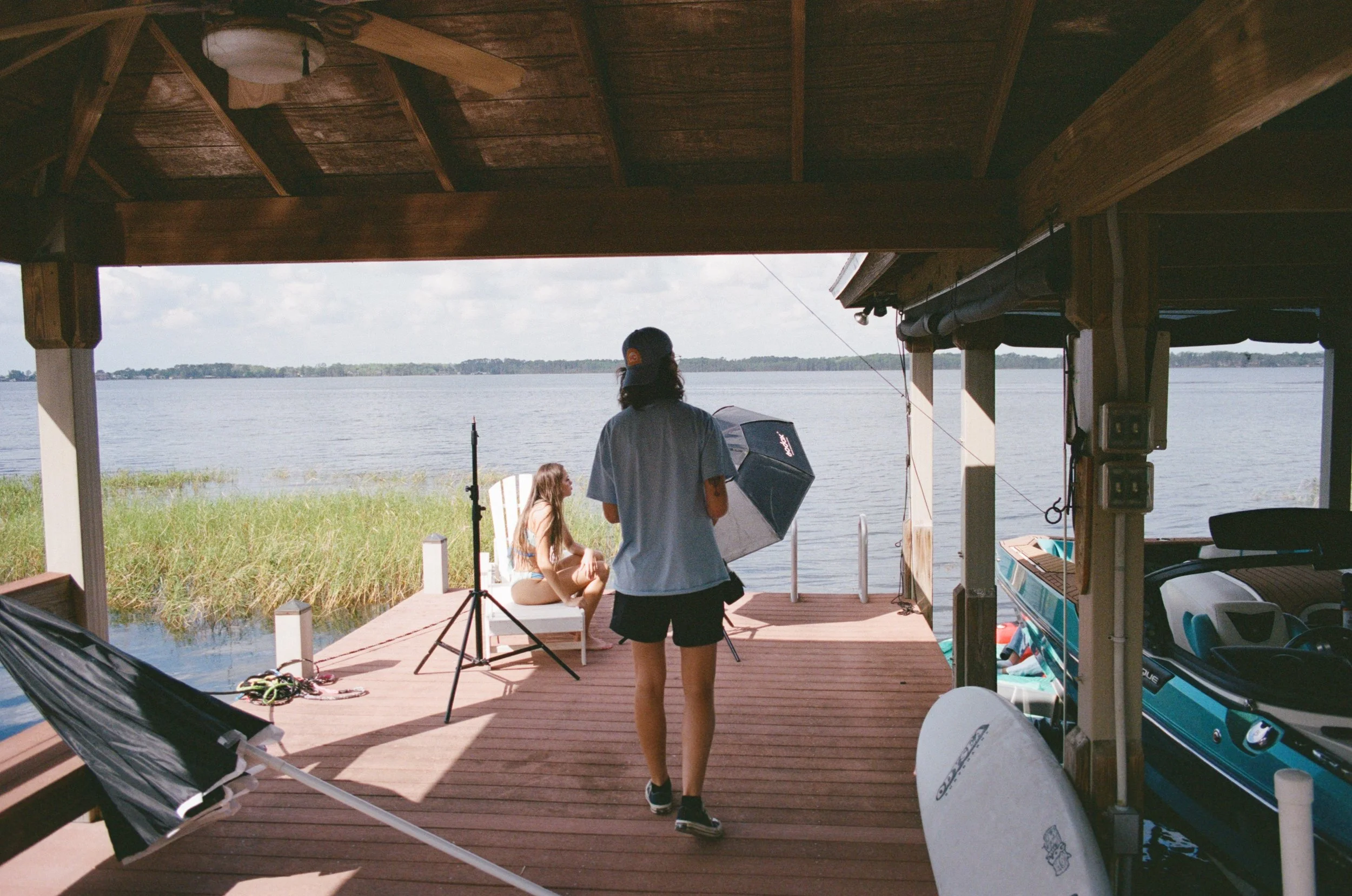 A woman sitting on a chair on a wooden dock by the water, while another woman stands nearby under a gazebo, holding a large black umbrella during a photoshoot setup by a lake, with watercraft and photography equipment around.