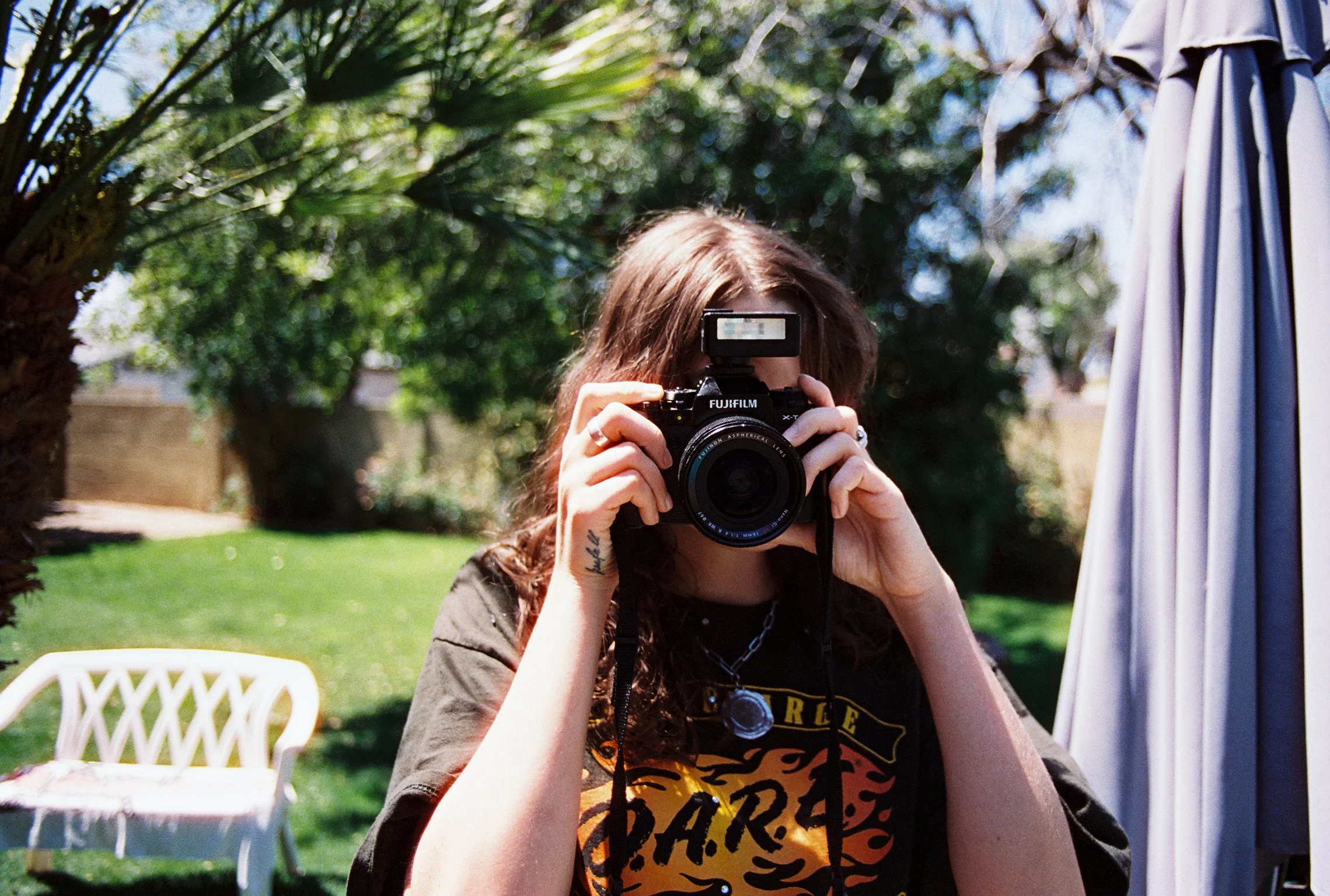Person with wavy hair holding a camera, standing outdoors on a sunny day, with greenery and a patio umbrella in the background.