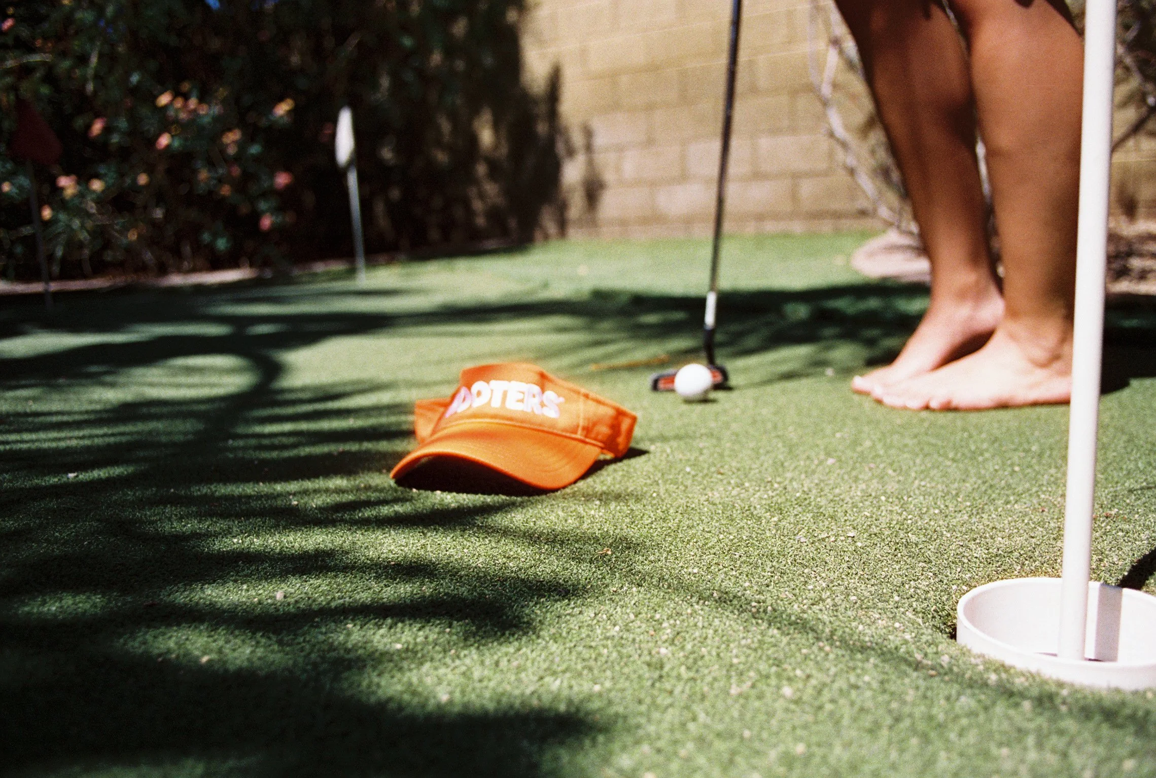 Close-up of a mini golf course with a green, orange Dippin' Dots hat, a golf ball, and a putter near a hole, with a person's bare feet and legs visible in the background.