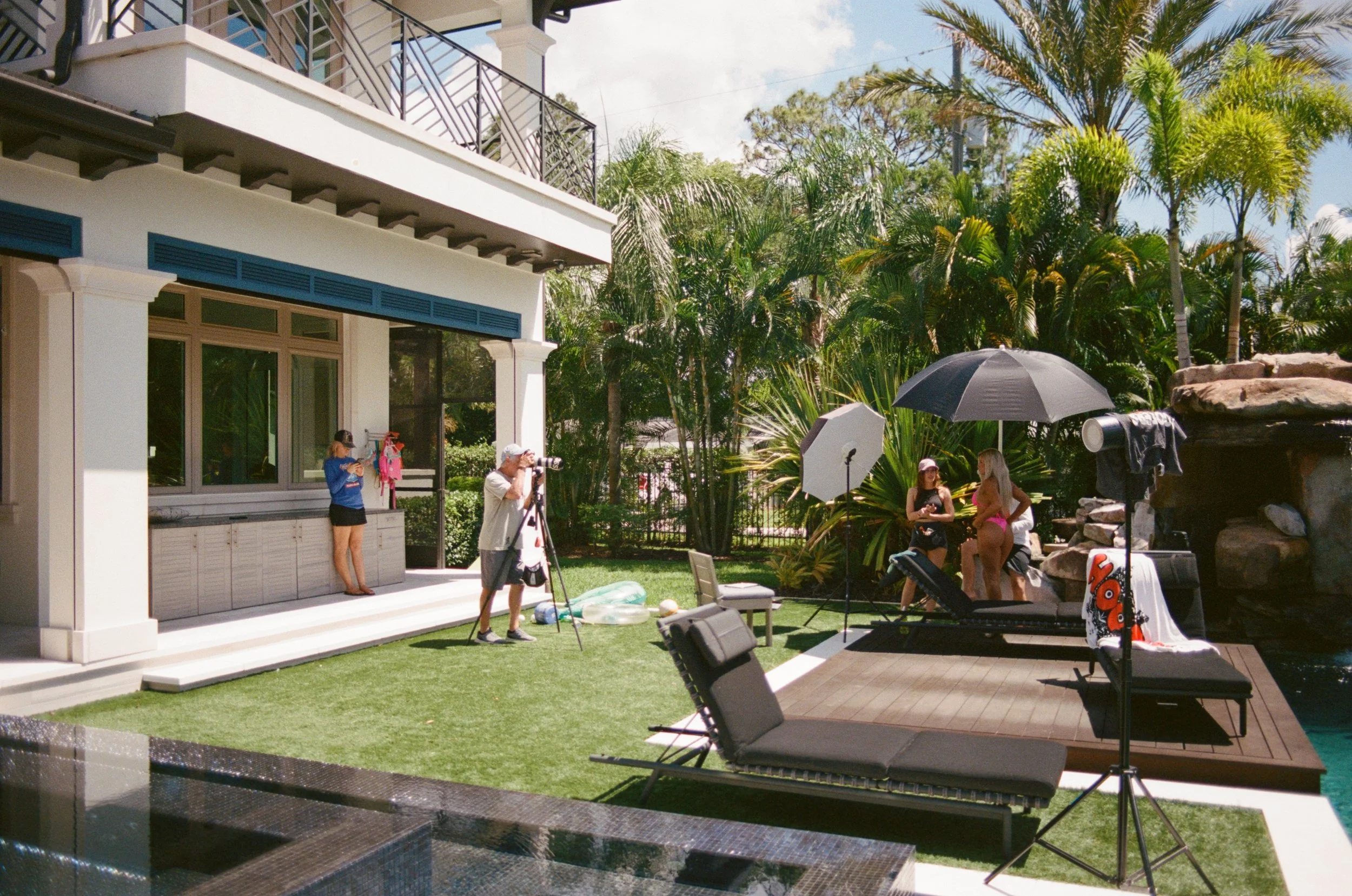 A backyard with a small pool, a patio area with lounge chairs, a large umbrella, and a photo shoot setup featuring a backdrop, professional lights, and models in swimsuits, with a photographer and crew adjusting equipment.