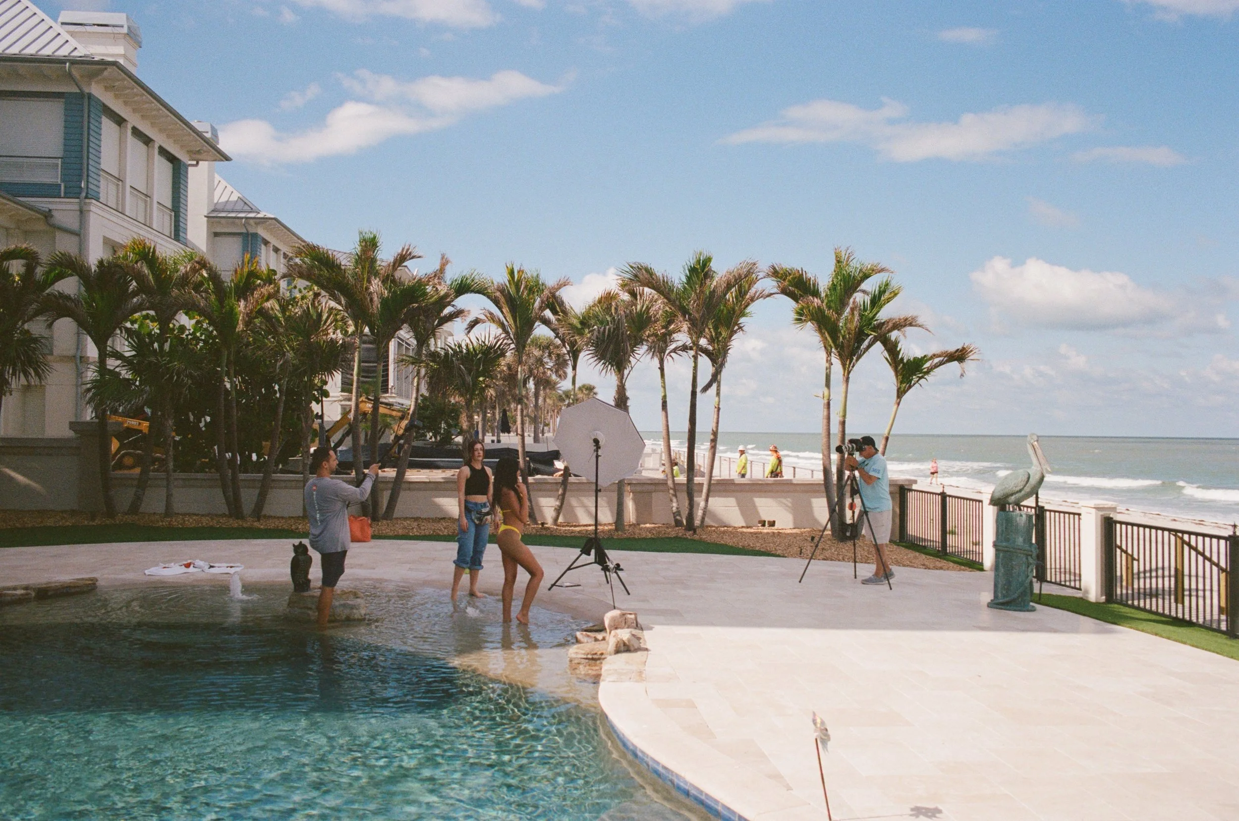 A photoshoot at a poolside near the beach with models, photographers, and crew, surrounded by palm trees and beachfront buildings.