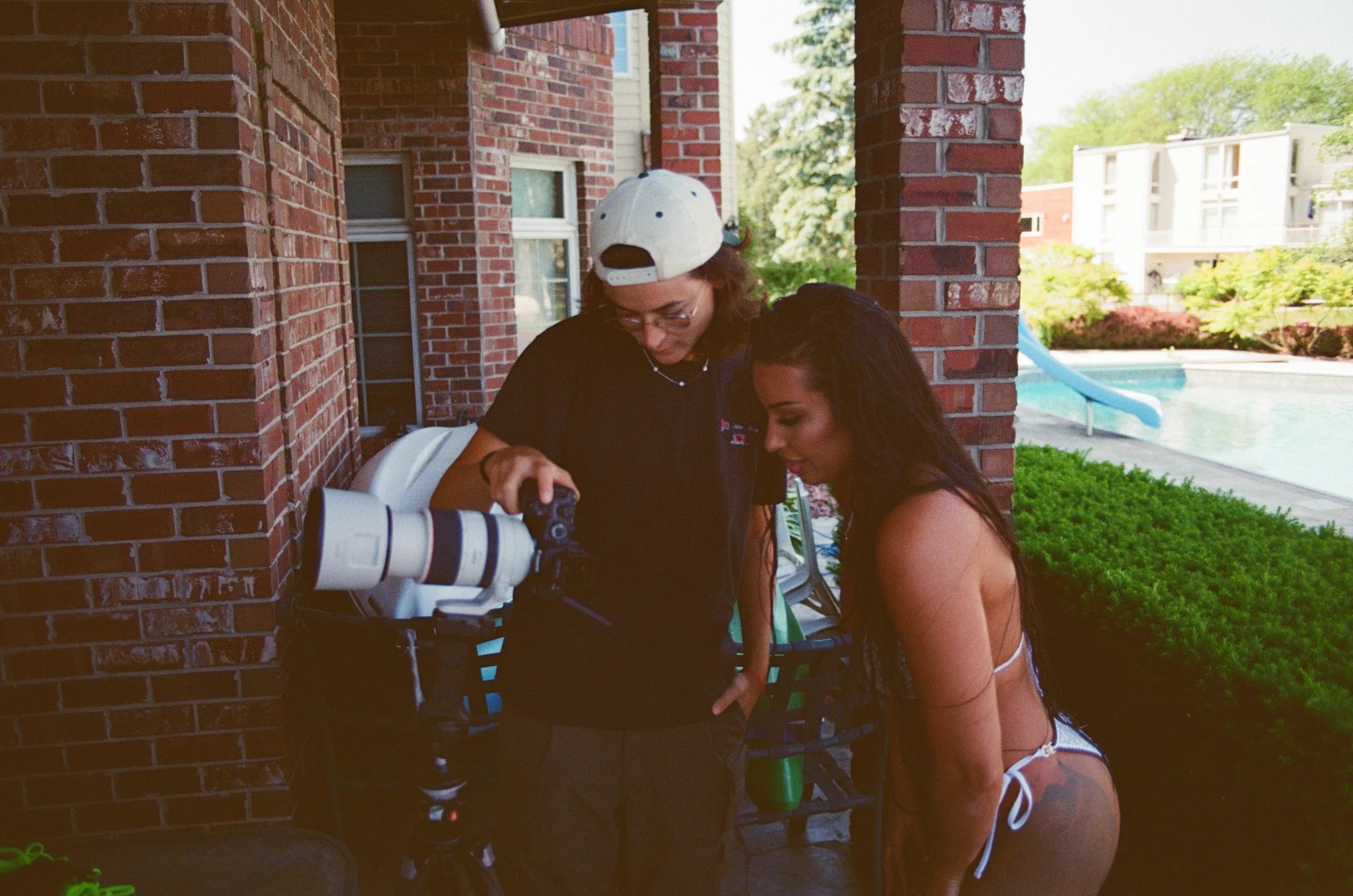 Two women looking at a camera with a large telephoto lens outdoors on a brick patio, with a swimming pool and slide in the background.