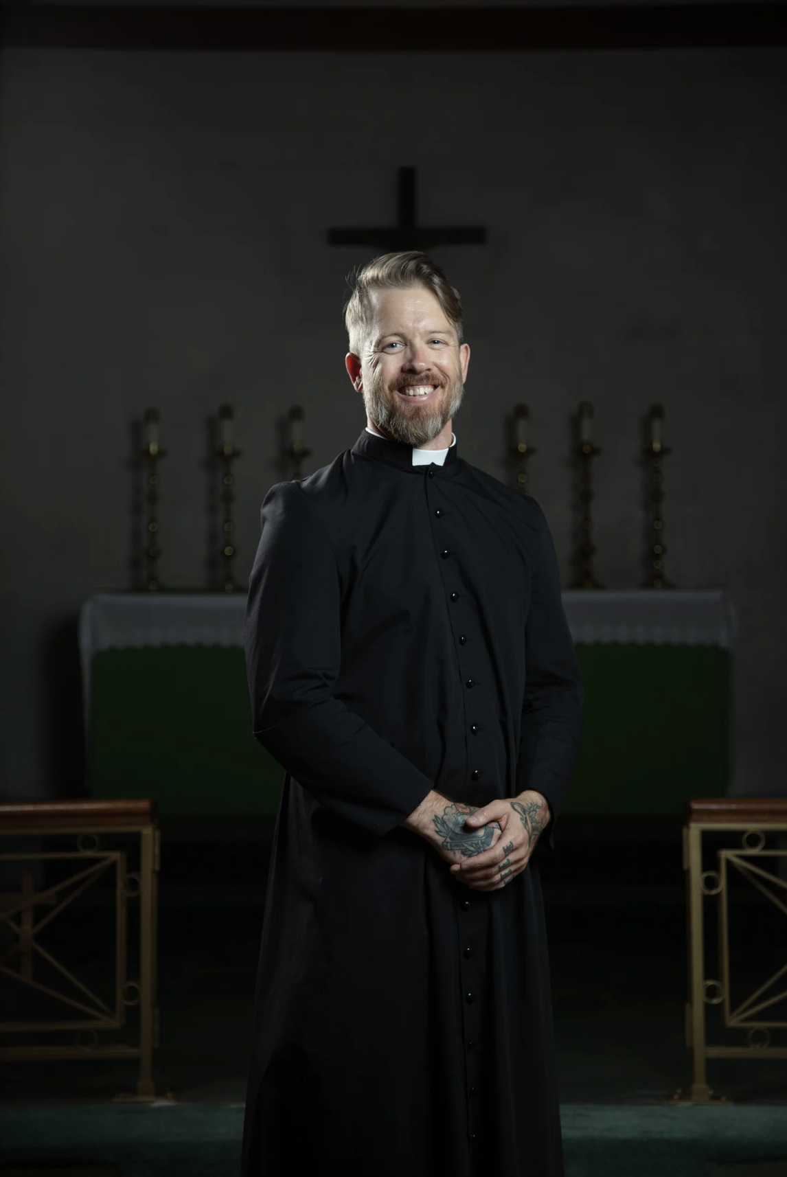 A smiling man dressed as a priest in a black robe standing inside a church, with a cross on the wall behind him.
