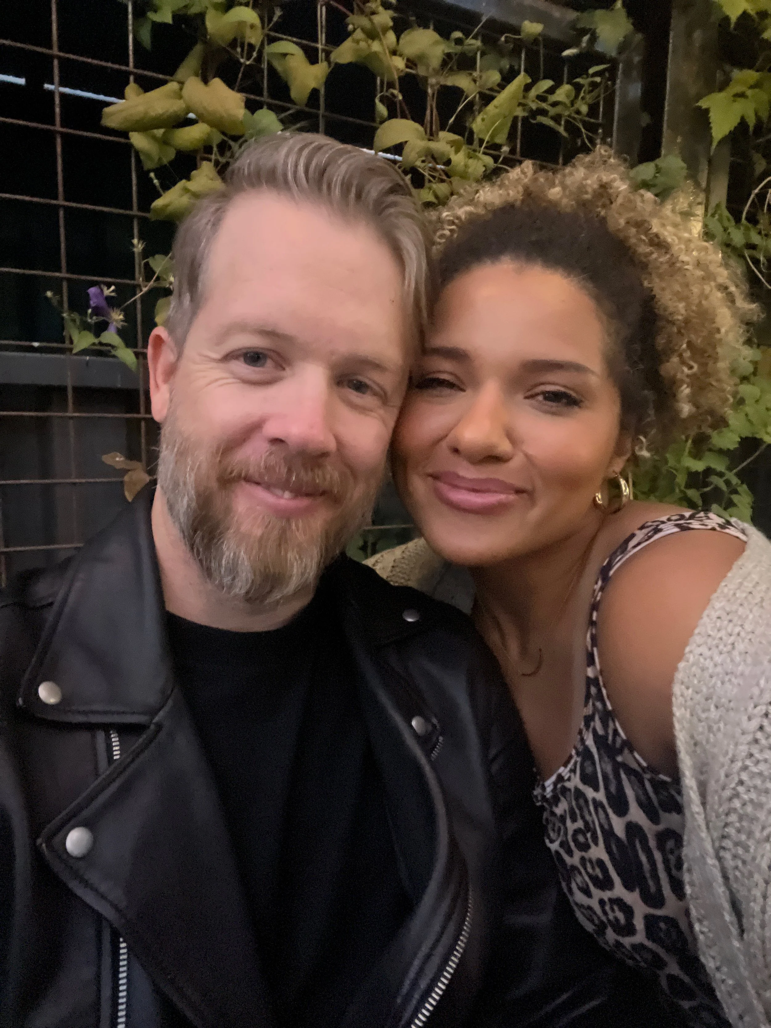 Close-up of a smiling man with a beard and a woman with curly hair posing together outdoors at night, with green foliage and a metal fence in the background.