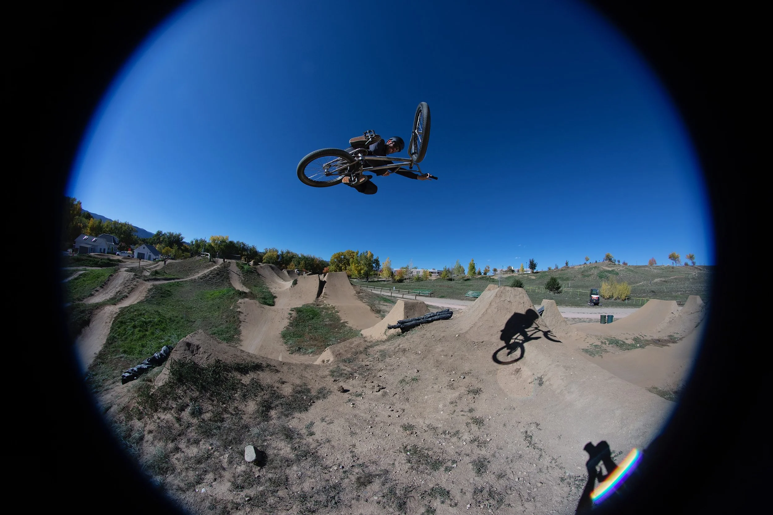 A person performing a BMX trick in mid-air at a dirt BMX pump track during daytime, with a clear blue sky and landscape in the background.