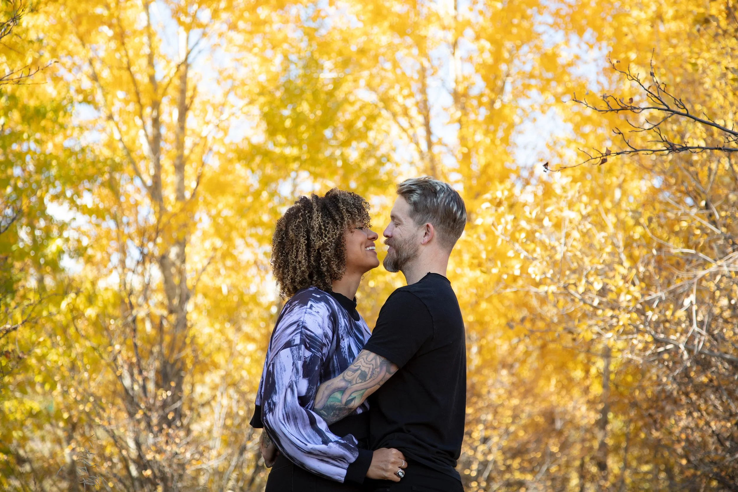 A couple standing close to each other in an autumn forest with yellow leaves, smiling and looking into each other's eyes.