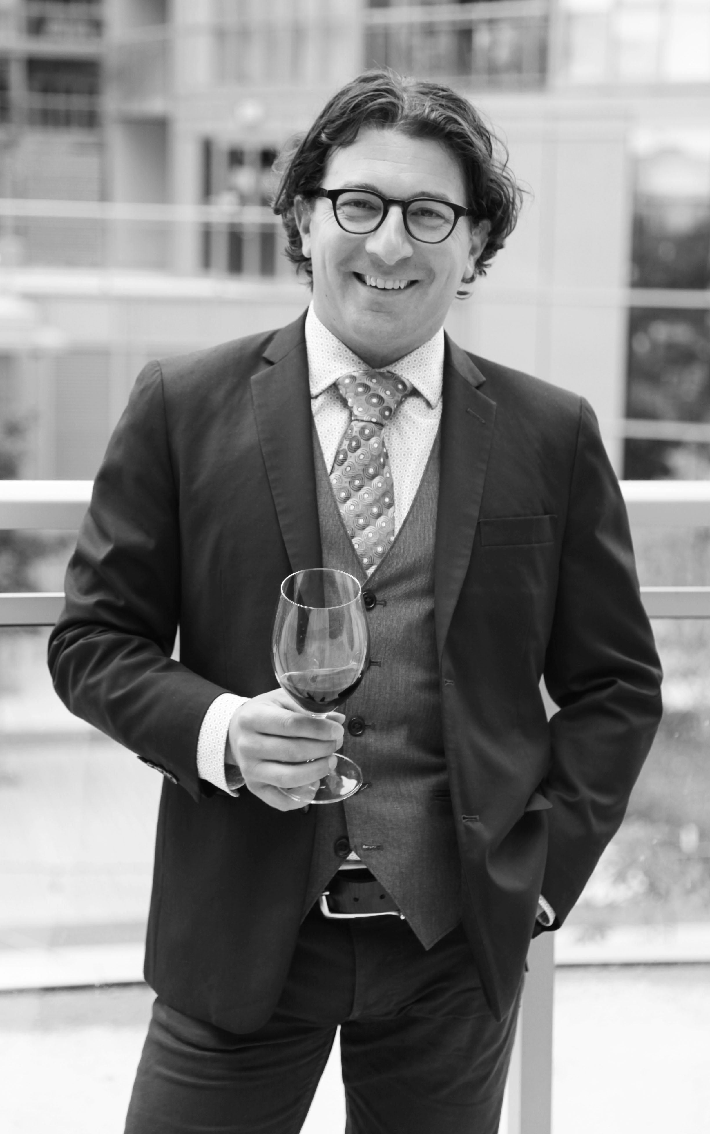 Black and white photo of a man in a suit holding a glass of red wine, smiling, outdoors with modern building in the background.