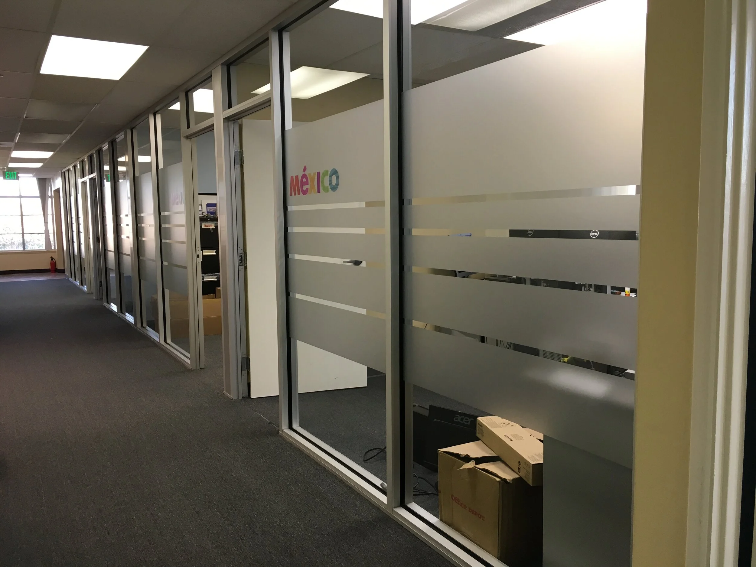 Empty office cubicles with glass partitions, some boxes, and office supplies, with a window and exit sign in the background.