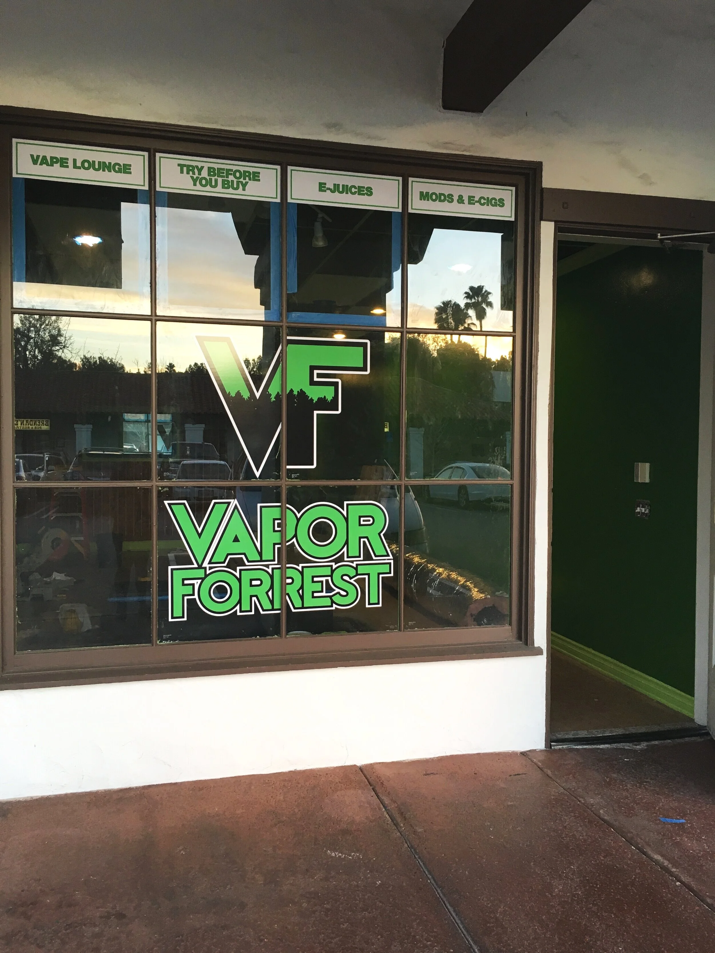Window display of Vapor Forest shop with green and black sign, including signs promoting vape lounge, try before you buy, E-Juices, mods and E-Cigs, and a logo with the letters V and F in green and black.