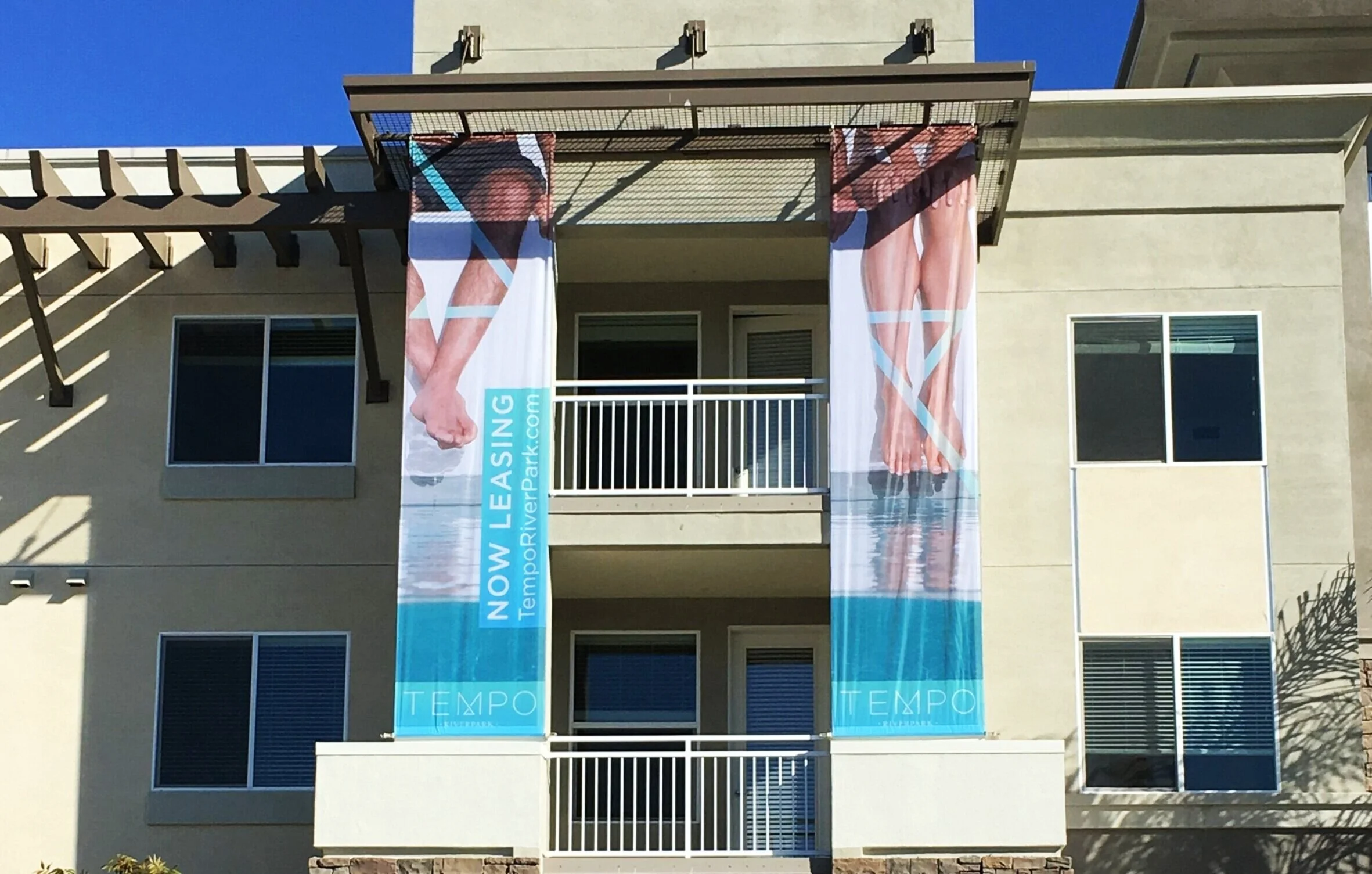 Three vertical banners hanging from the balcony of a modern building with large windows. The banners have images of a woman in swimwear and the text 'NOW LEASING' and 'TEMPO'. The building appears to be under sunlight with clear blue sky in the backg