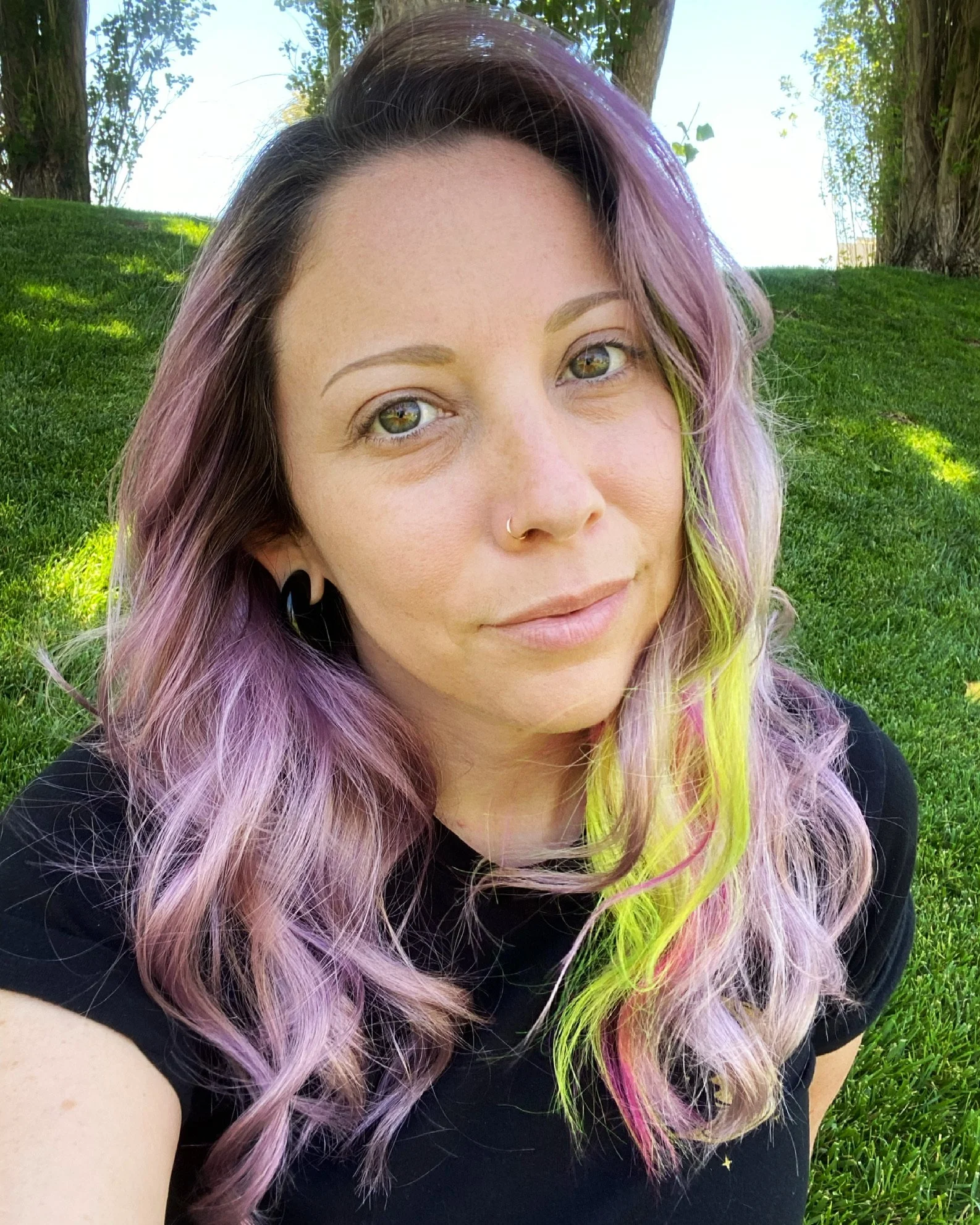 A young woman with multicolored hair, including shades of purple, pink, yellow, and green, is outdoors on a grass field with trees in the background. She has a slight smile, light-colored eyes, and a nose ring.