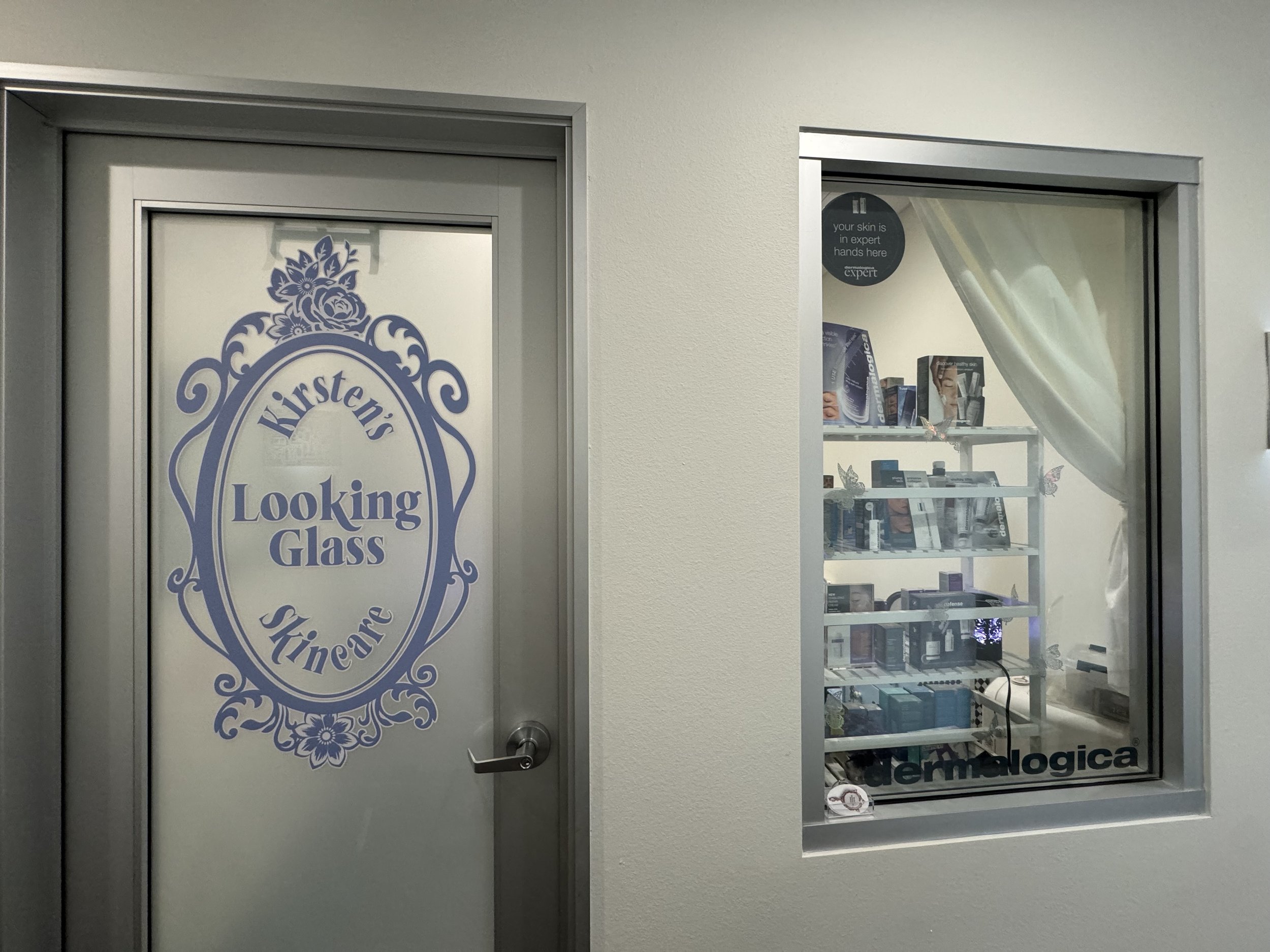A door with a sign for a hearing care center and a window with skincare products inside, next to a white wall.