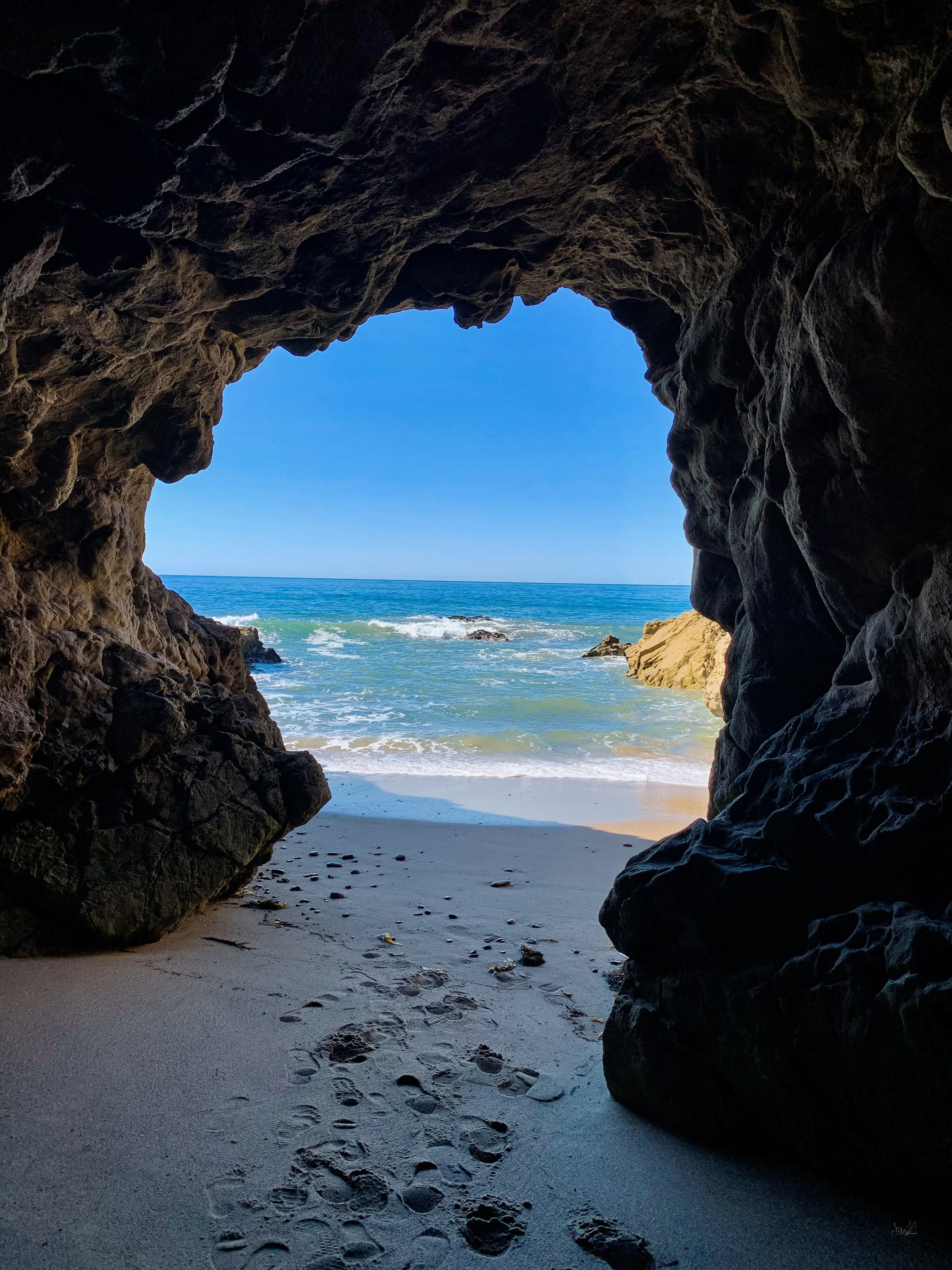View from inside a rocky cave looking out to a sandy beach and the ocean with a clear blue sky.