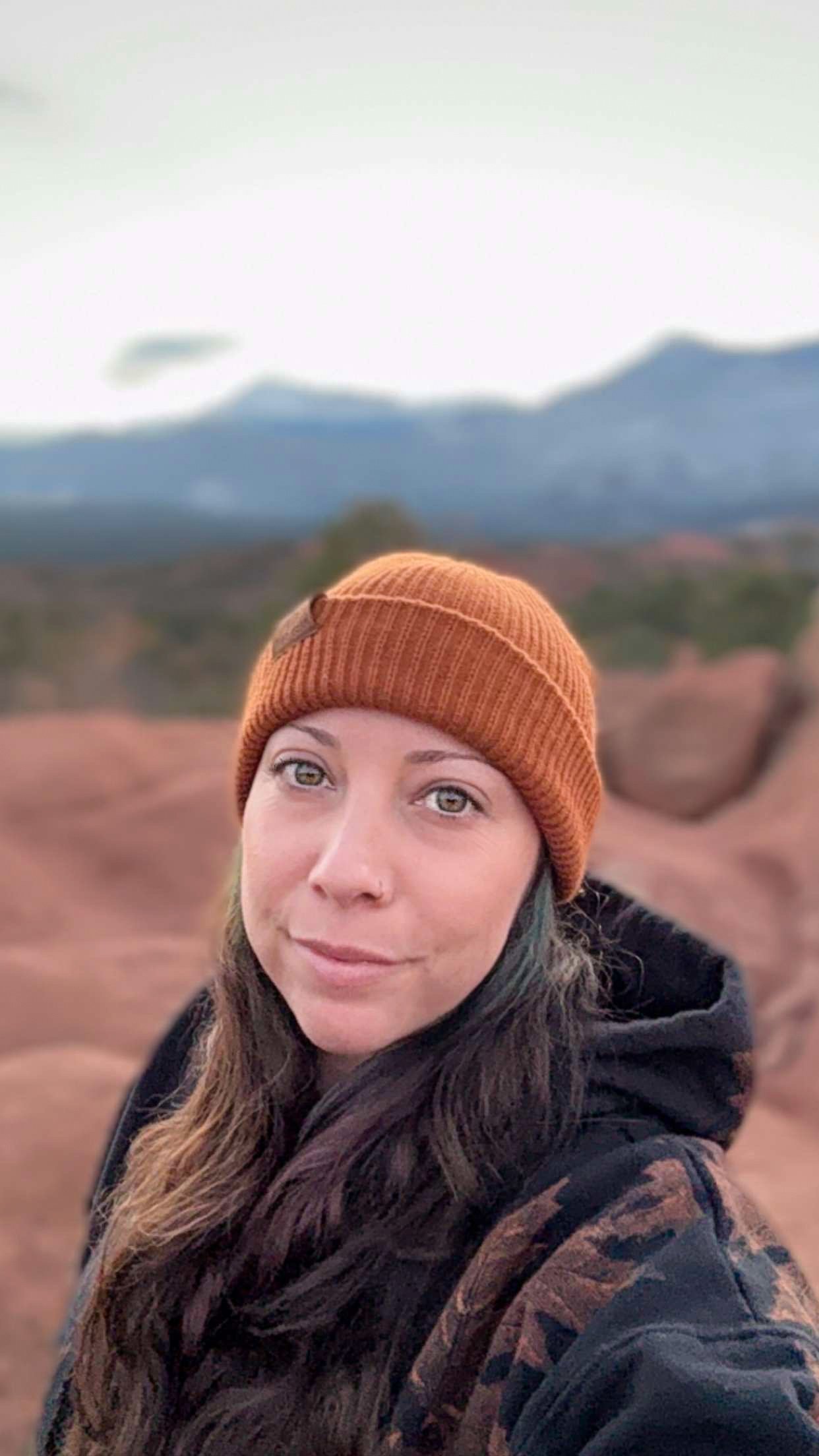 Close-up of a woman with long brown hair wearing an orange knit beanie and a camouflage jacket, standing outdoors with mountains and cloudy sky in the background.