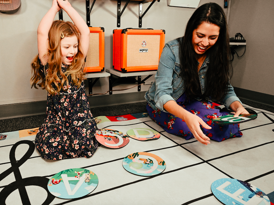 Child and adult playing musical games with letter cards on treble clef mat in room with orange amplifiers.