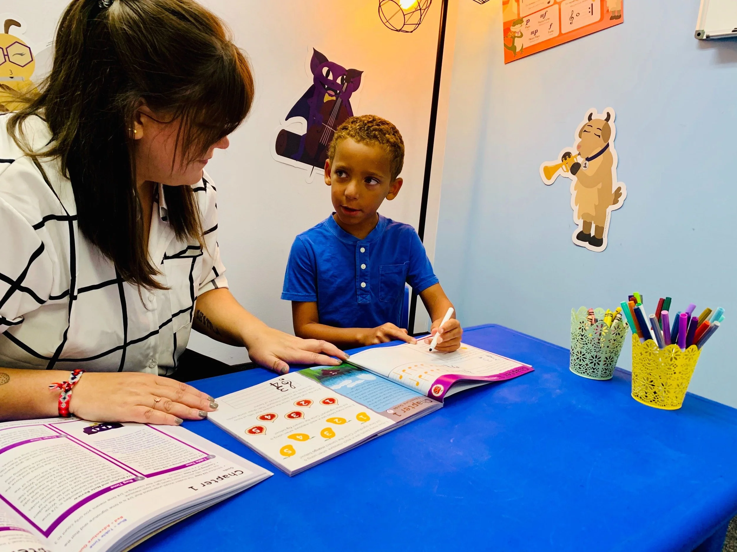 A teacher and a young student sit at a blue table with open workbooks and colored pens. The wall has animal decorations and educational posters.