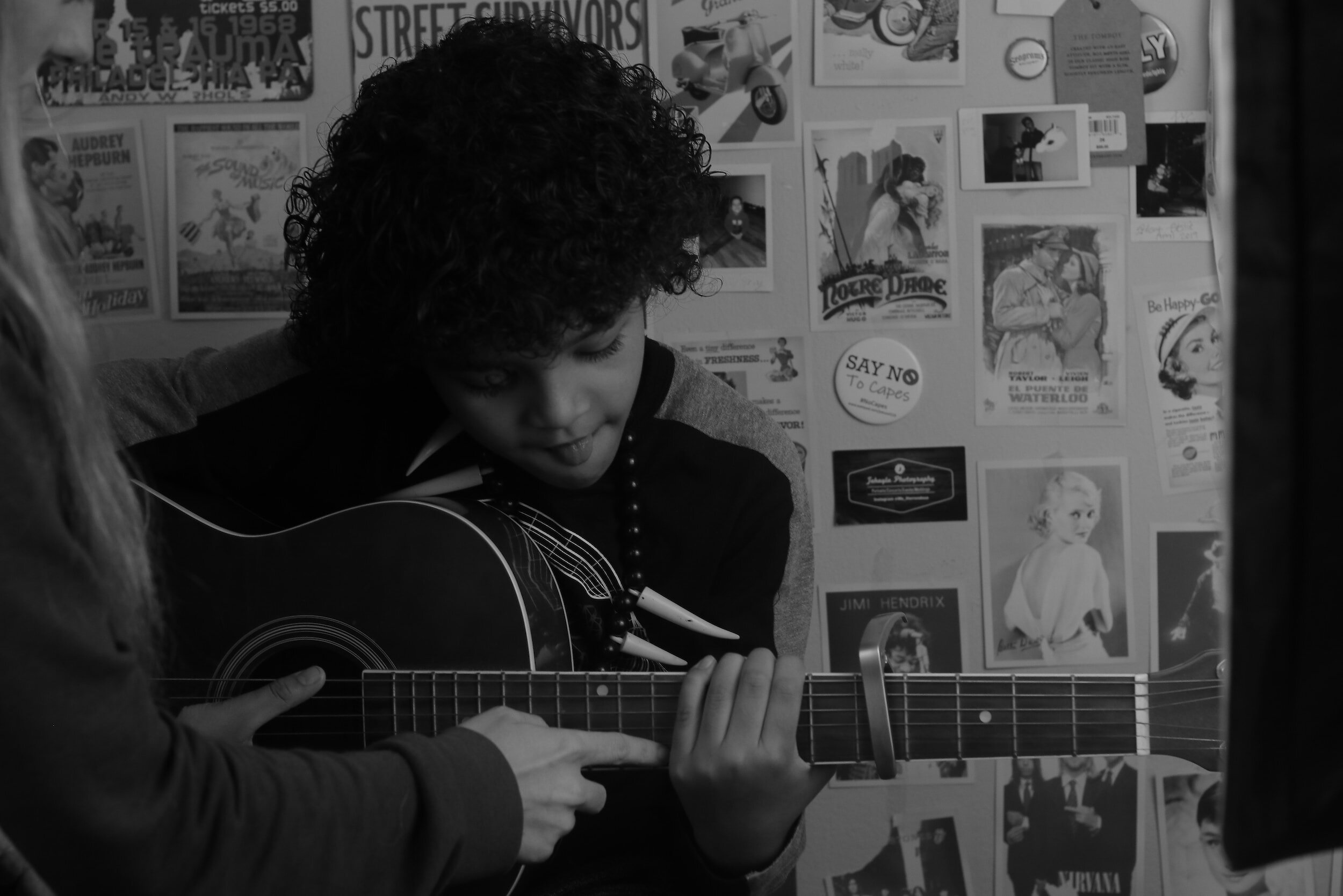 Child learning to play an acoustic guitar with guidance from an adult, surrounded by vintage posters on the wall.