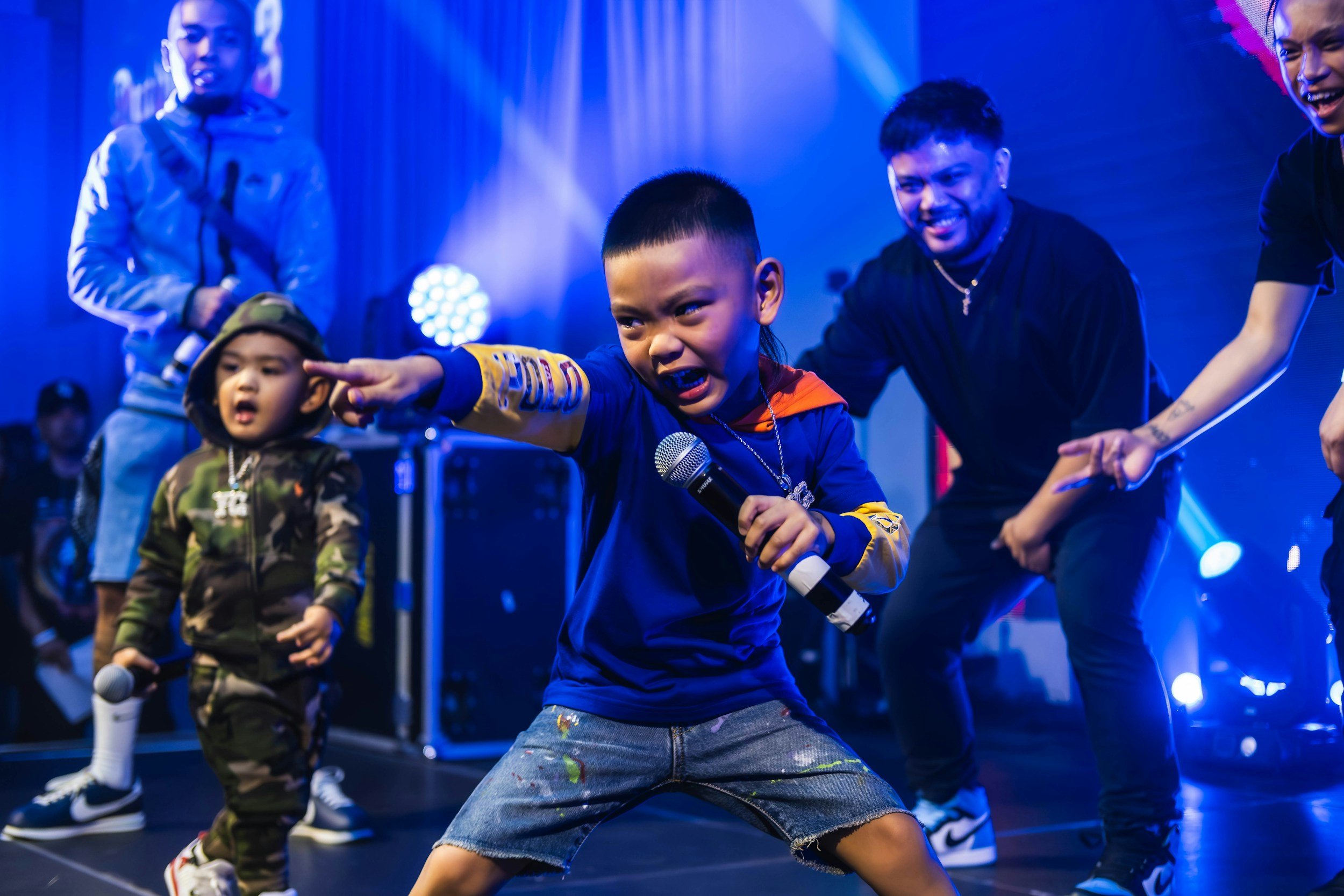 Three young boys performing energetically on stage with microphones, under blue stage lights.