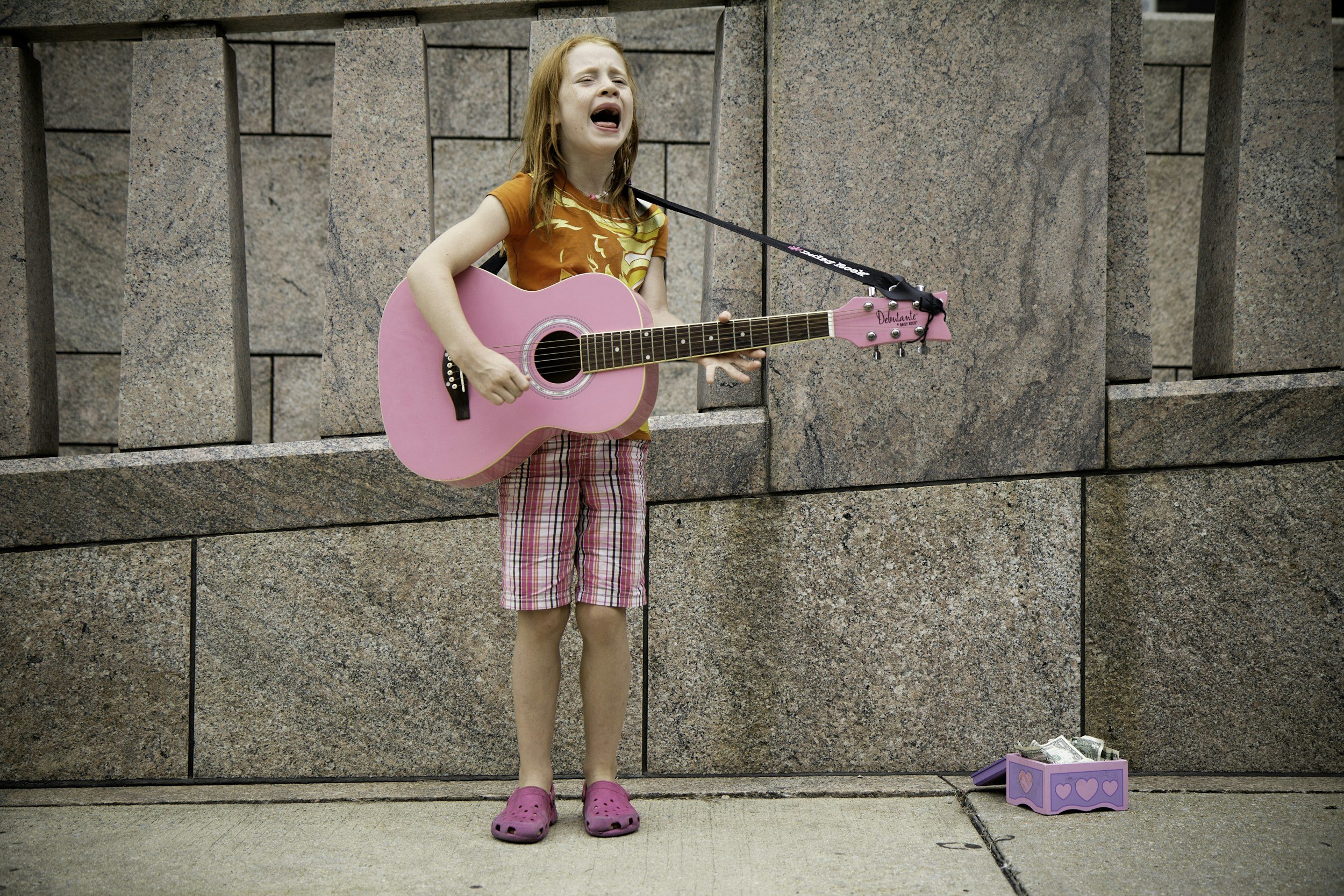 Young girl playing a pink guitar and singing on a sidewalk, wearing plaid shorts and a t-shirt, with a tip box on the ground.