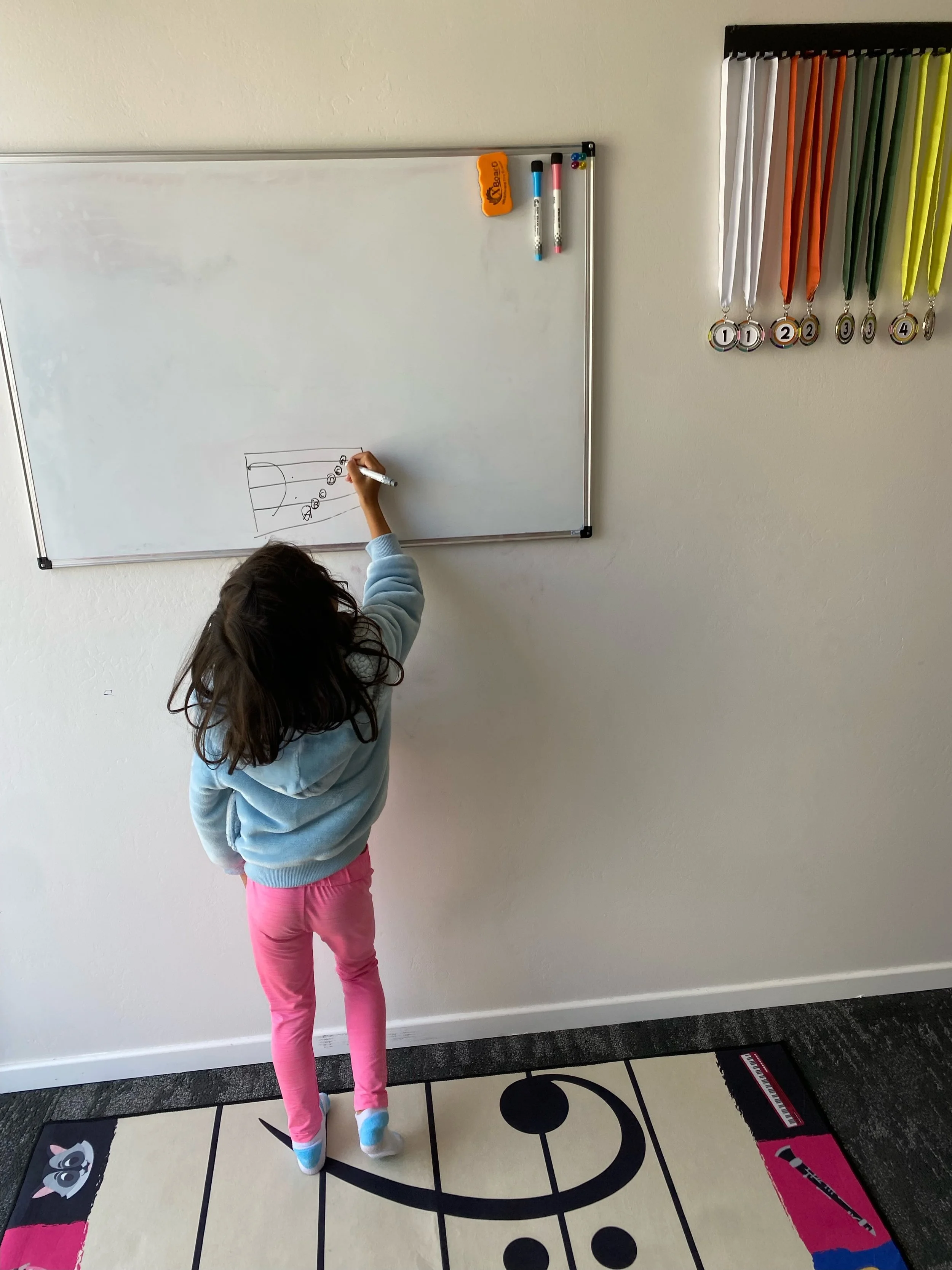 A child in colorful clothing draws on a whiteboard, with various medals hanging on the wall beside the board.