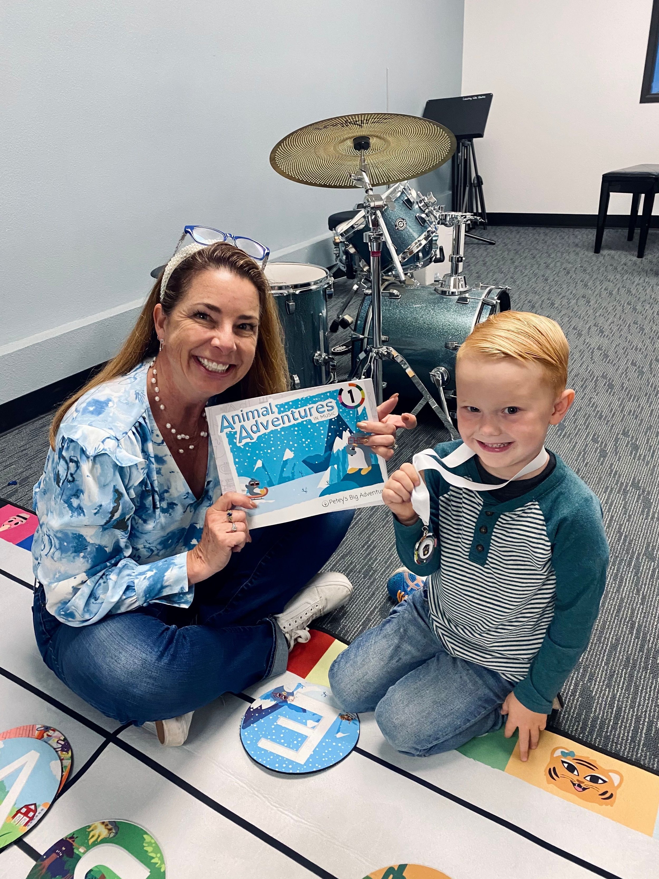 Woman and child holding a children's book titled "Animal Adventures" with a drum set in the background. The child is holding a medal. They are sitting on a colorful mat in a music room.