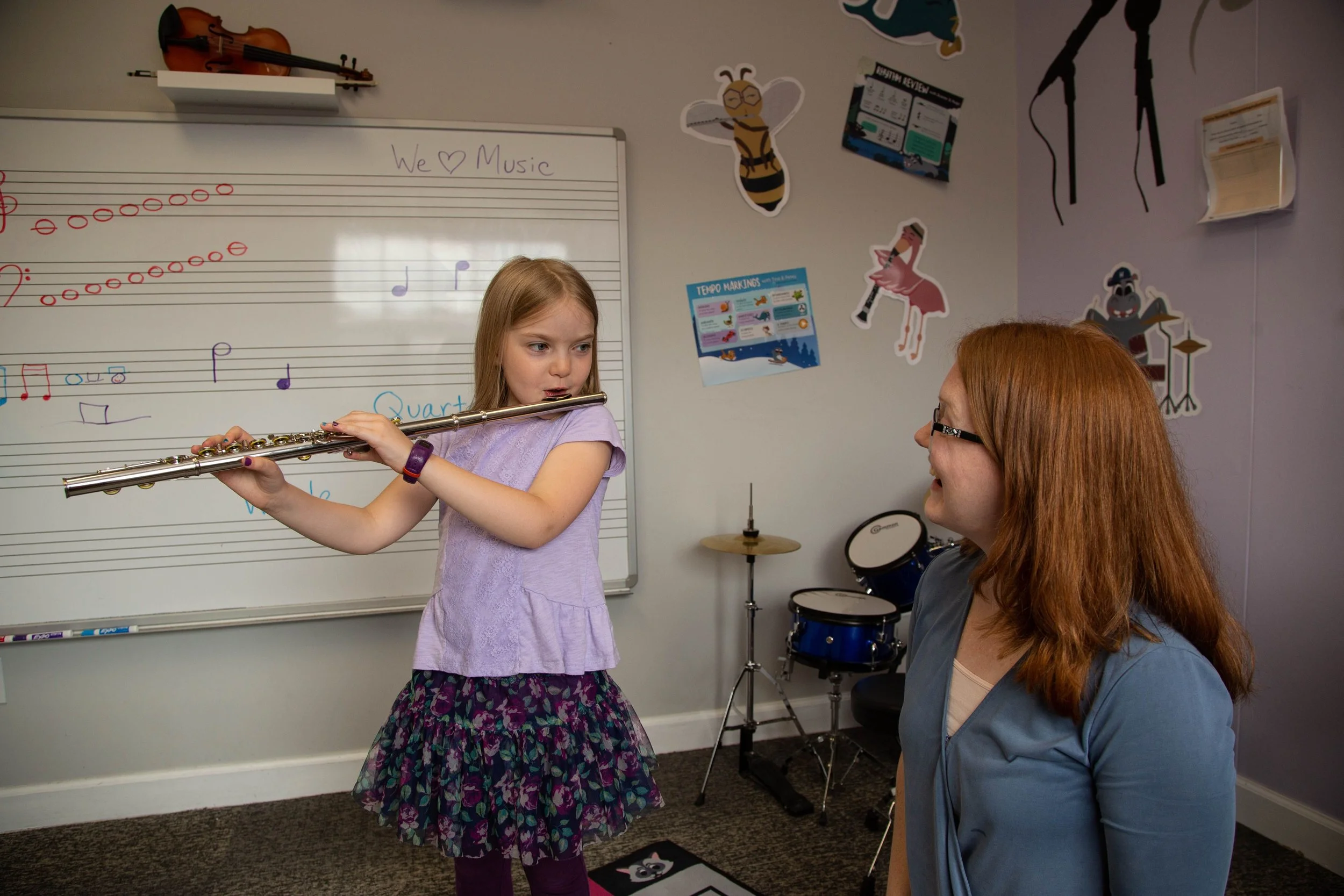 Young girl playing the flute in a music classroom with a teacher watching. A whiteboard with musical notes is visible, along with wall decorations and a small drum set in the background.