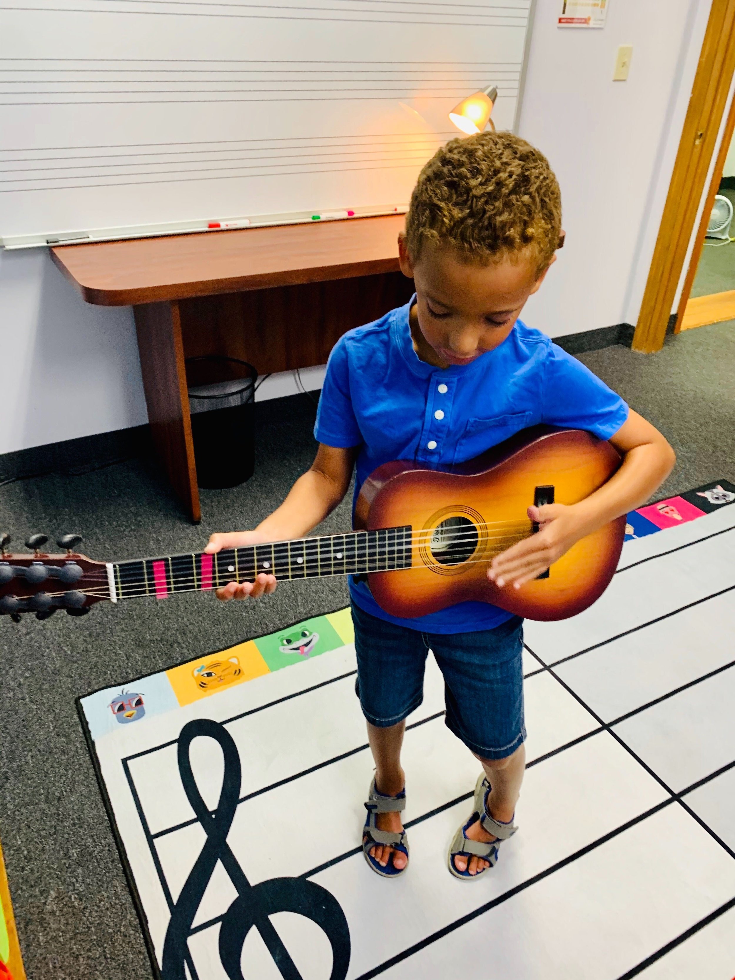 Child in blue shirt playing acoustic guitar in a music classroom with staff lines on the floor.