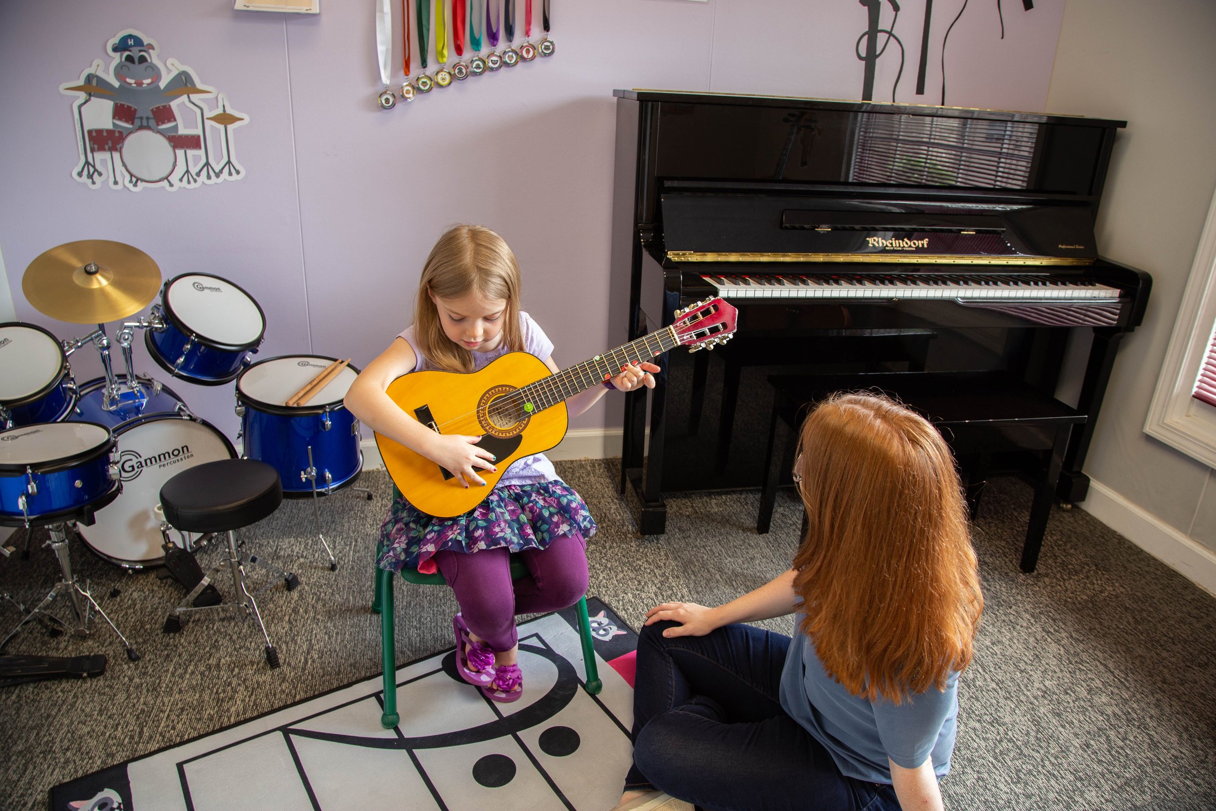 Child playing a guitar with an adult watching, alongside a piano and drum set in a music room.