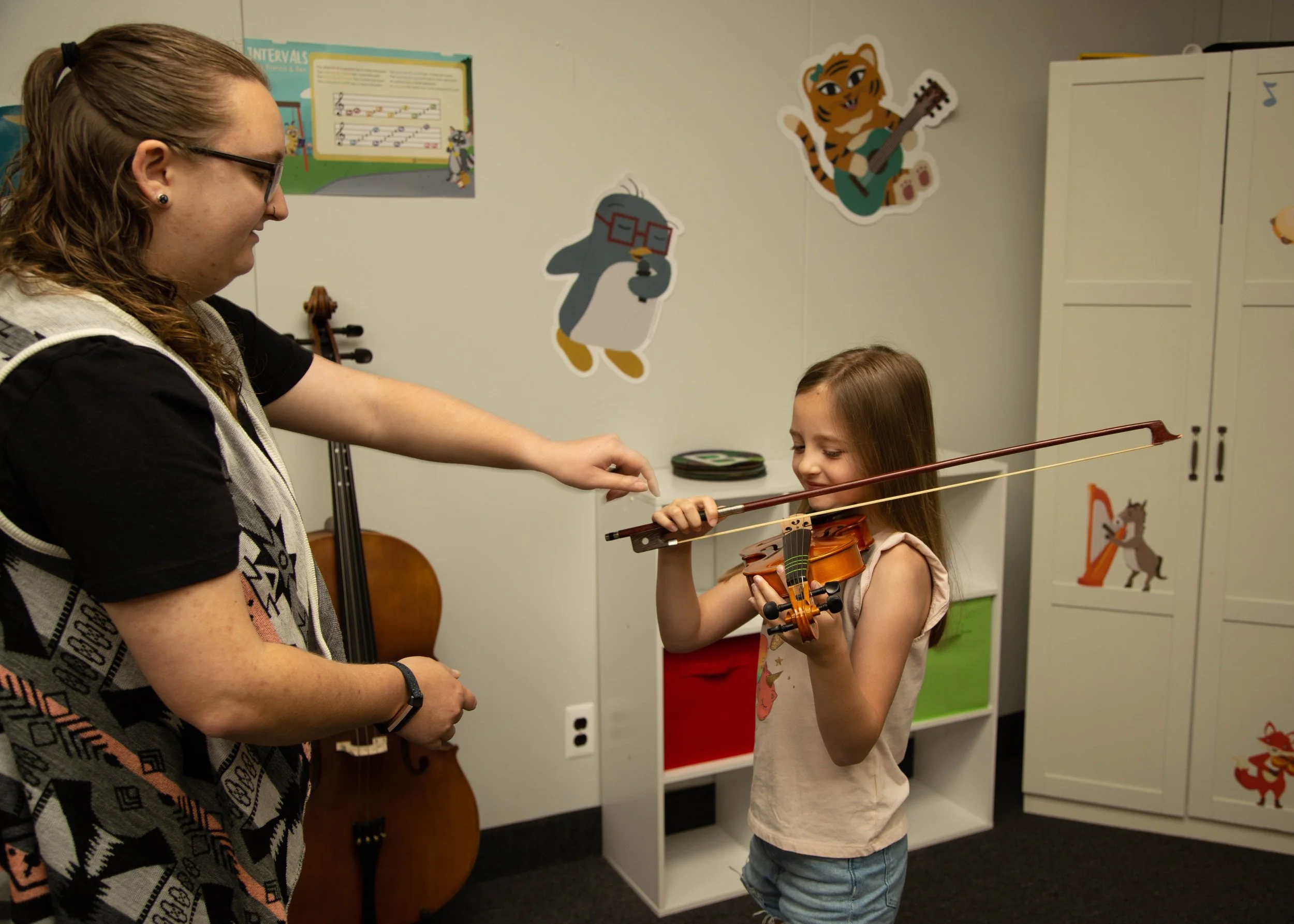 A young girl playing a violin with guidance from an adult in a music classroom decorated with cartoon animal posters and musical instruments.