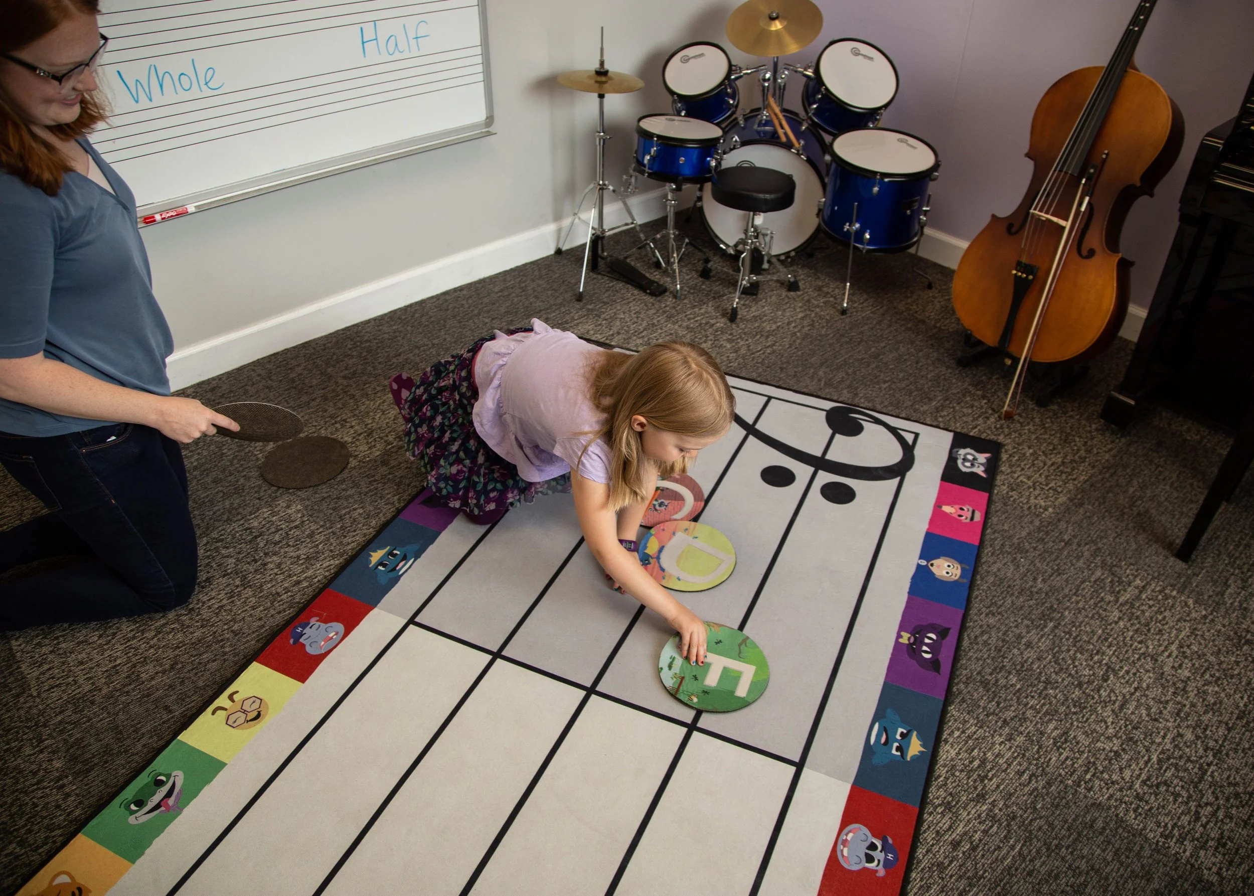 Child arranging musical notes on a large staff rug in a music classroom, with drum set and double bass in the background. A teacher is kneeling nearby, observing.