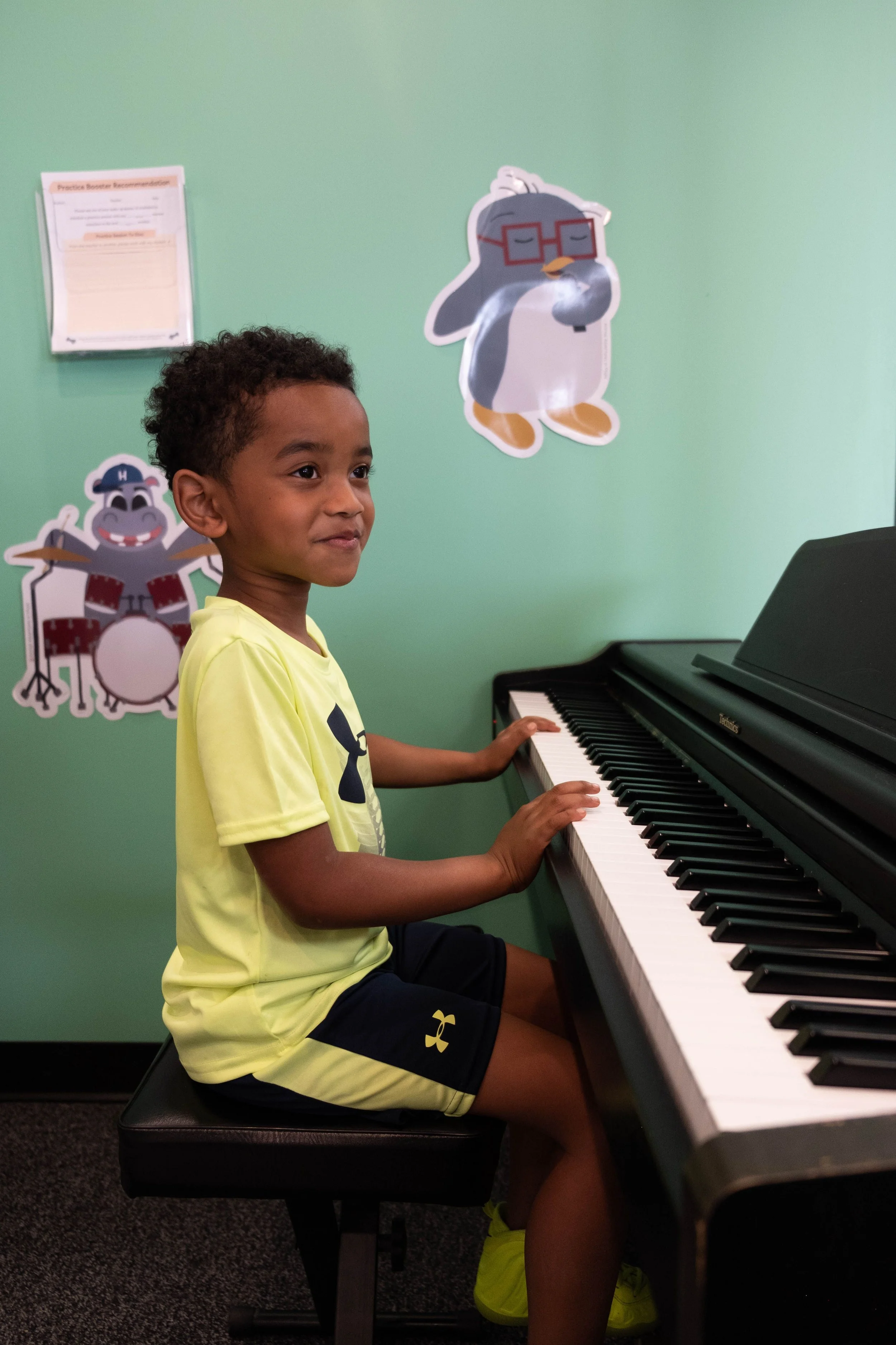 Child in a bright yellow outfit playing a piano in a room with wall decorations of cartoon animals, including a penguin and a hippopotamus.