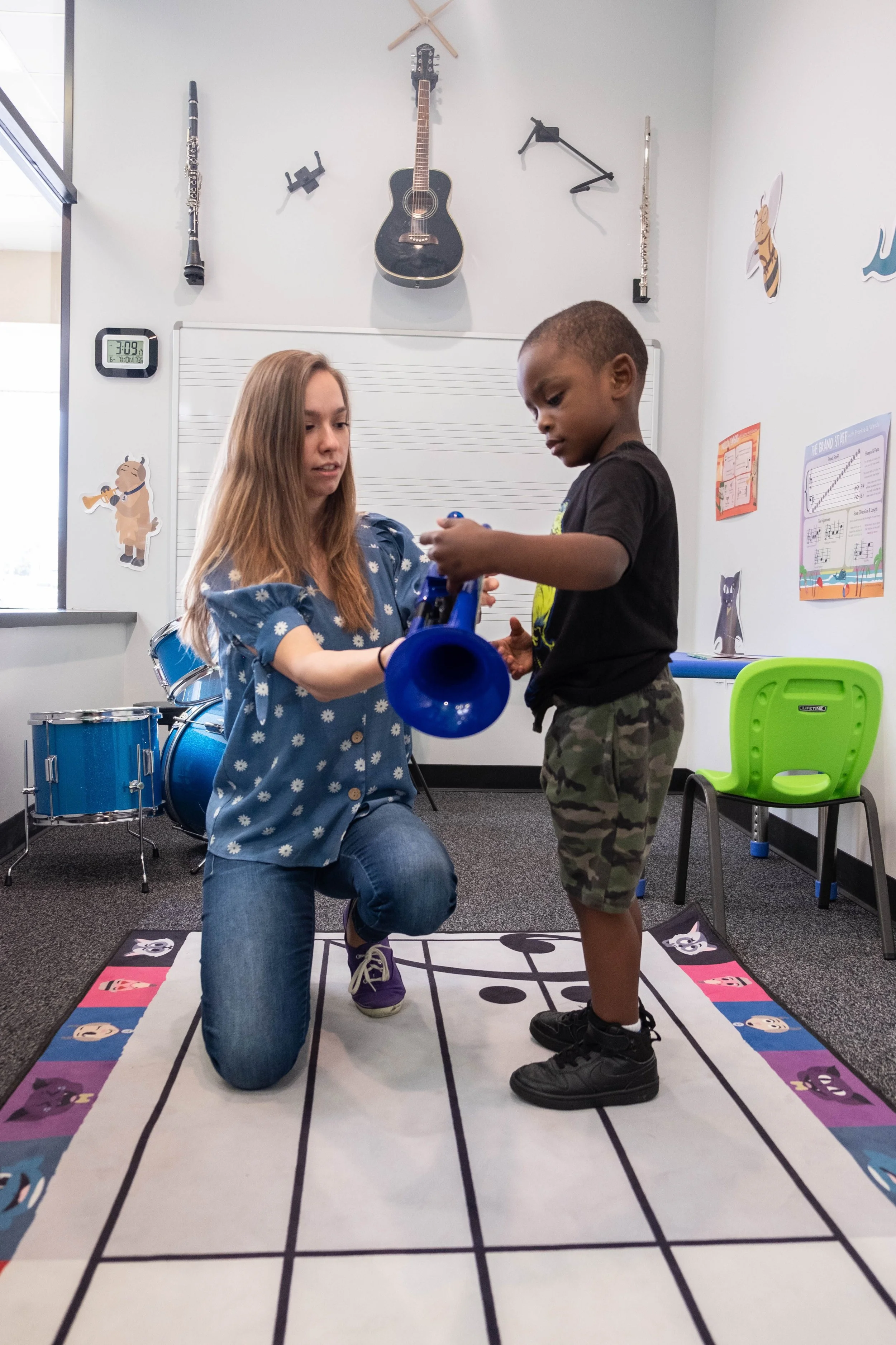 A woman kneels on a music note-themed carpet, assisting a young boy holding a blue trumpet in a music classroom environment. The room has musical instruments like a guitar and clarinet mounted on the wall, along with musical decor and educational posters. A drum set is visible in the background.