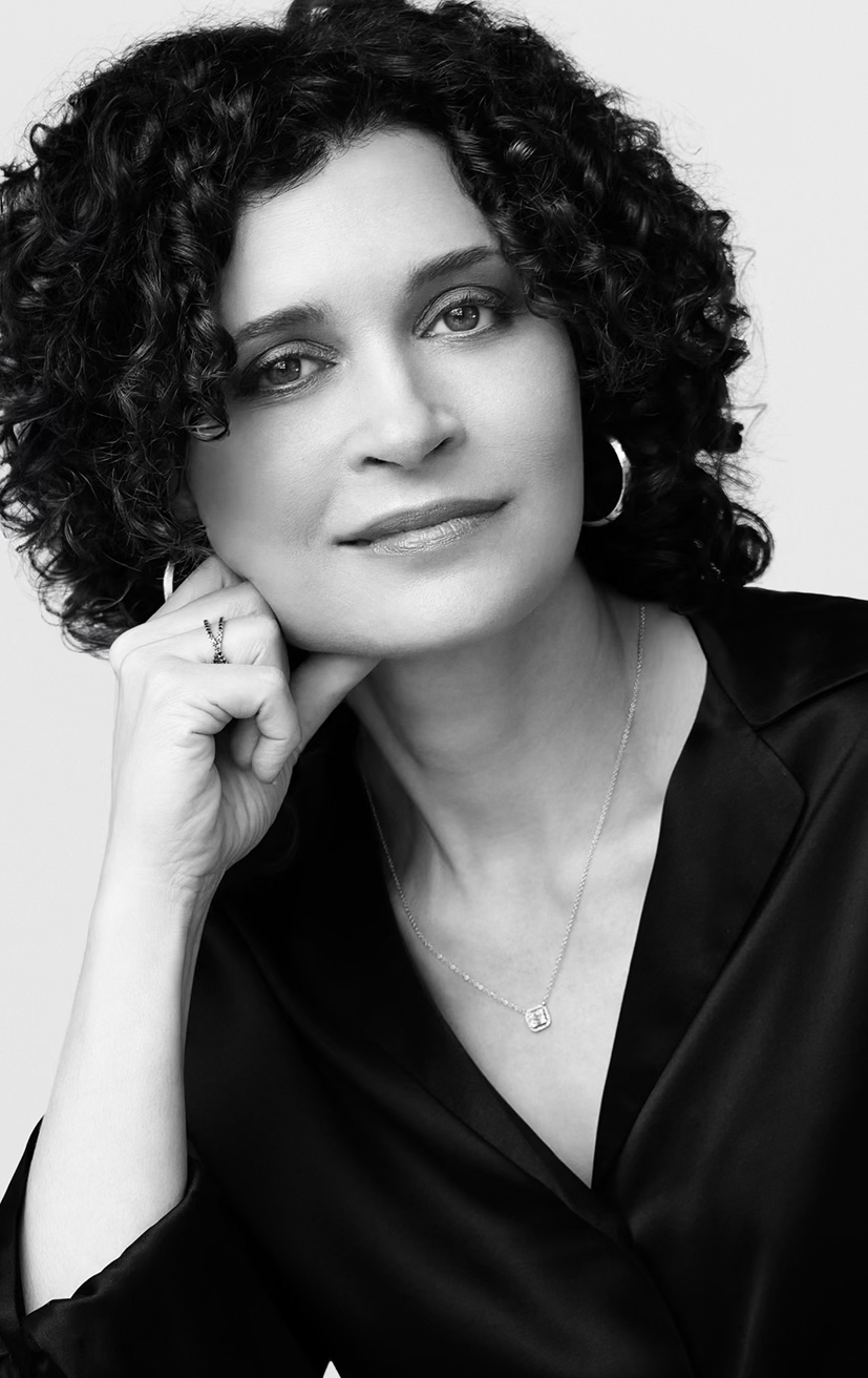 Black and white portrait of a woman with curly hair, wearing a black top, hoop earrings, a necklace, and a ring, resting her chin on her hand.