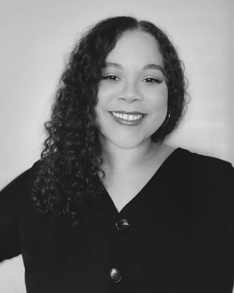 Black and white portrait of a woman with curly hair, smiling, wearing a black top with three buttons, against a plain background.