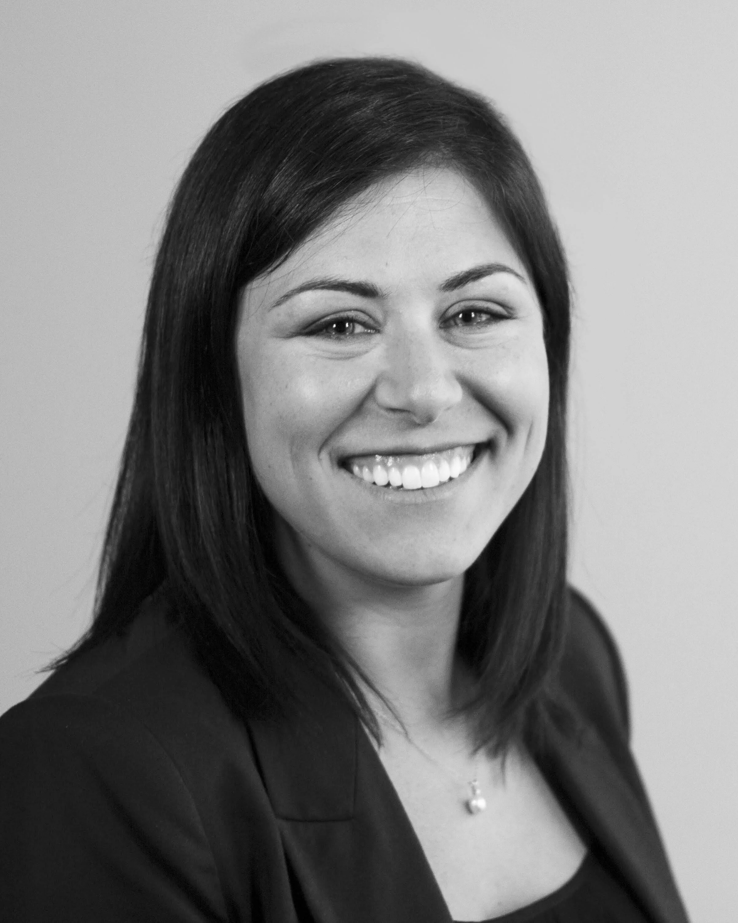 A woman with straight dark hair smiling, wearing a dark jacket and a necklace with a small pendant, with a plain background.