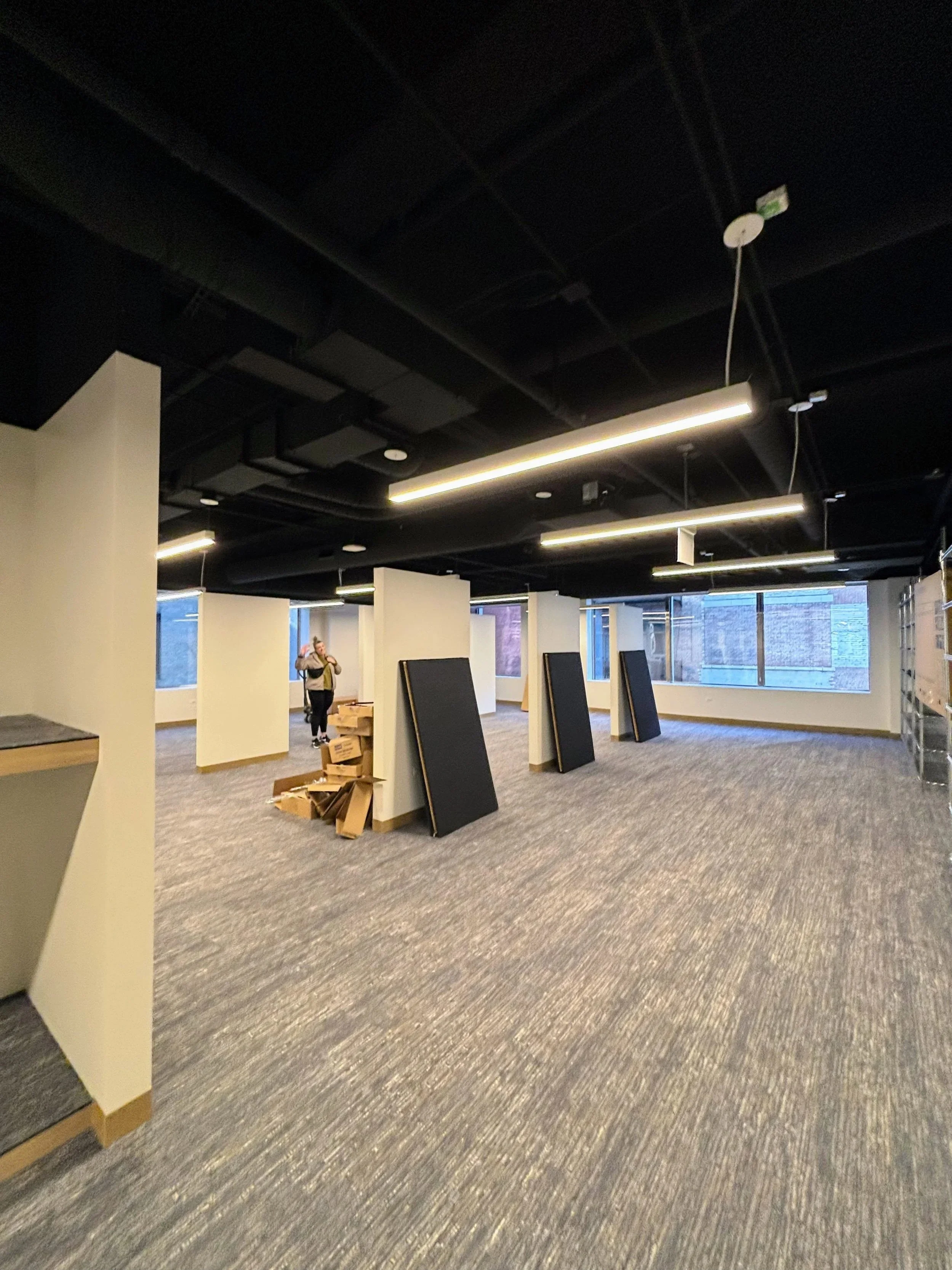 An indoor space with a high black ceiling and fluorescent lighting, beige walls, large windows, and some furniture being assembled or moved, including boxed items and display boards.