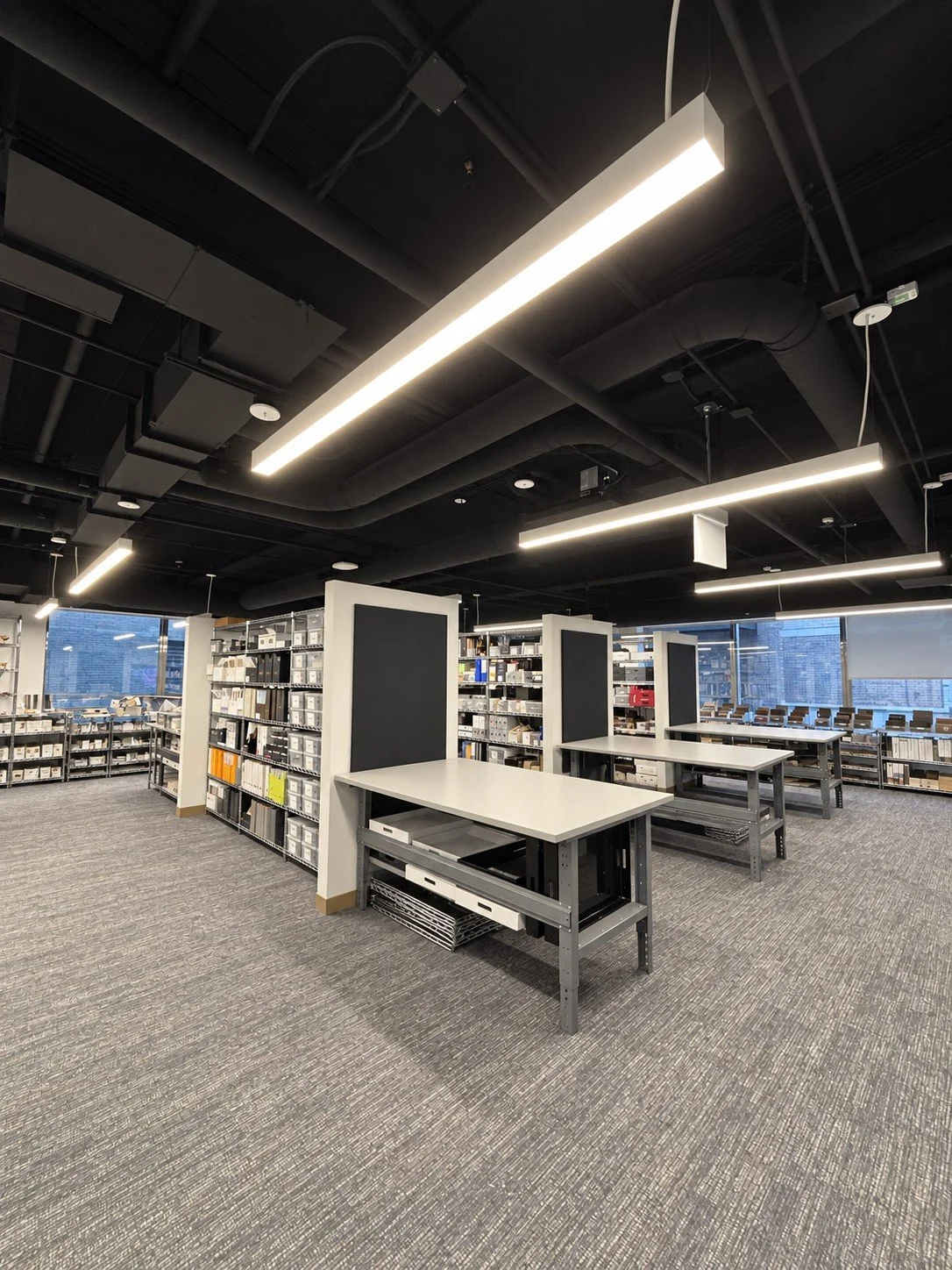 Modern library or office space with shelves of books or files, tables with dividers, and large windows.