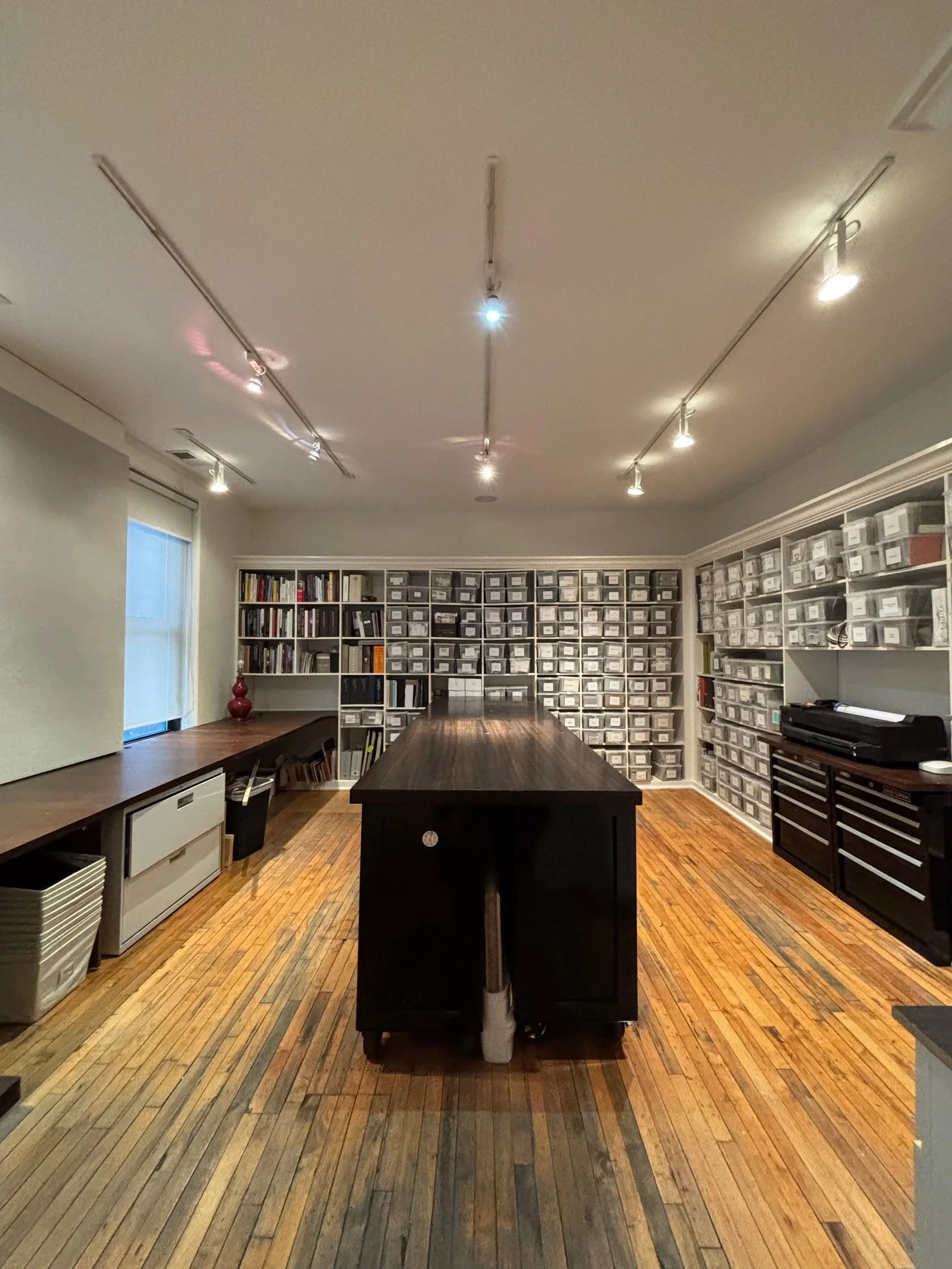 A room with a wooden floor, a large dark wooden table in the center, bookshelves and storage bins filled with various items against the far wall, a window with blinds on the left, and track lighting on the ceiling illuminating the room.