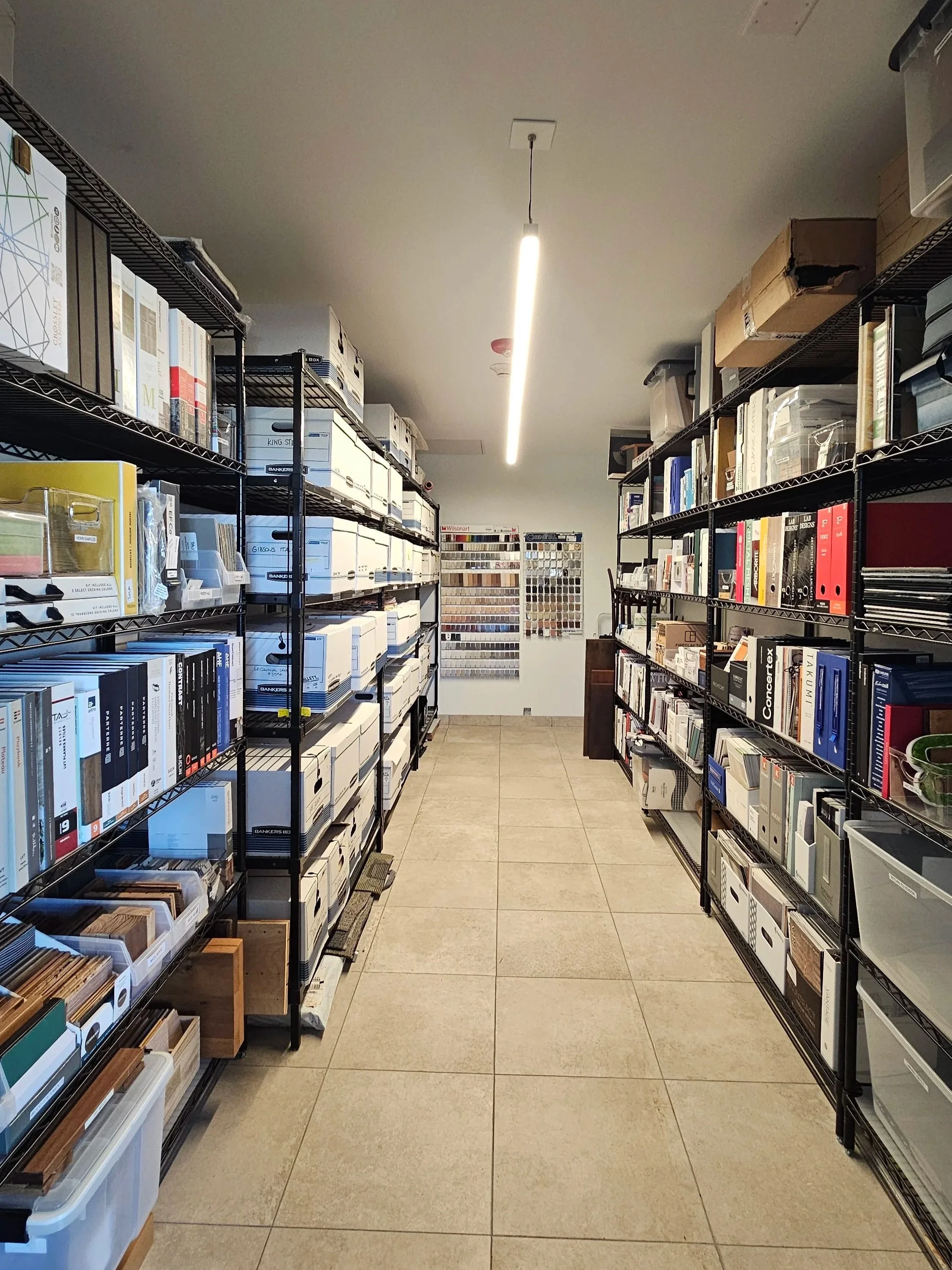 A store aisle with metal shelves stocked with books, binders, and storage boxes. The shelves are black wire, and the floor is beige tile. Fluorescent lighting runs along the ceiling.