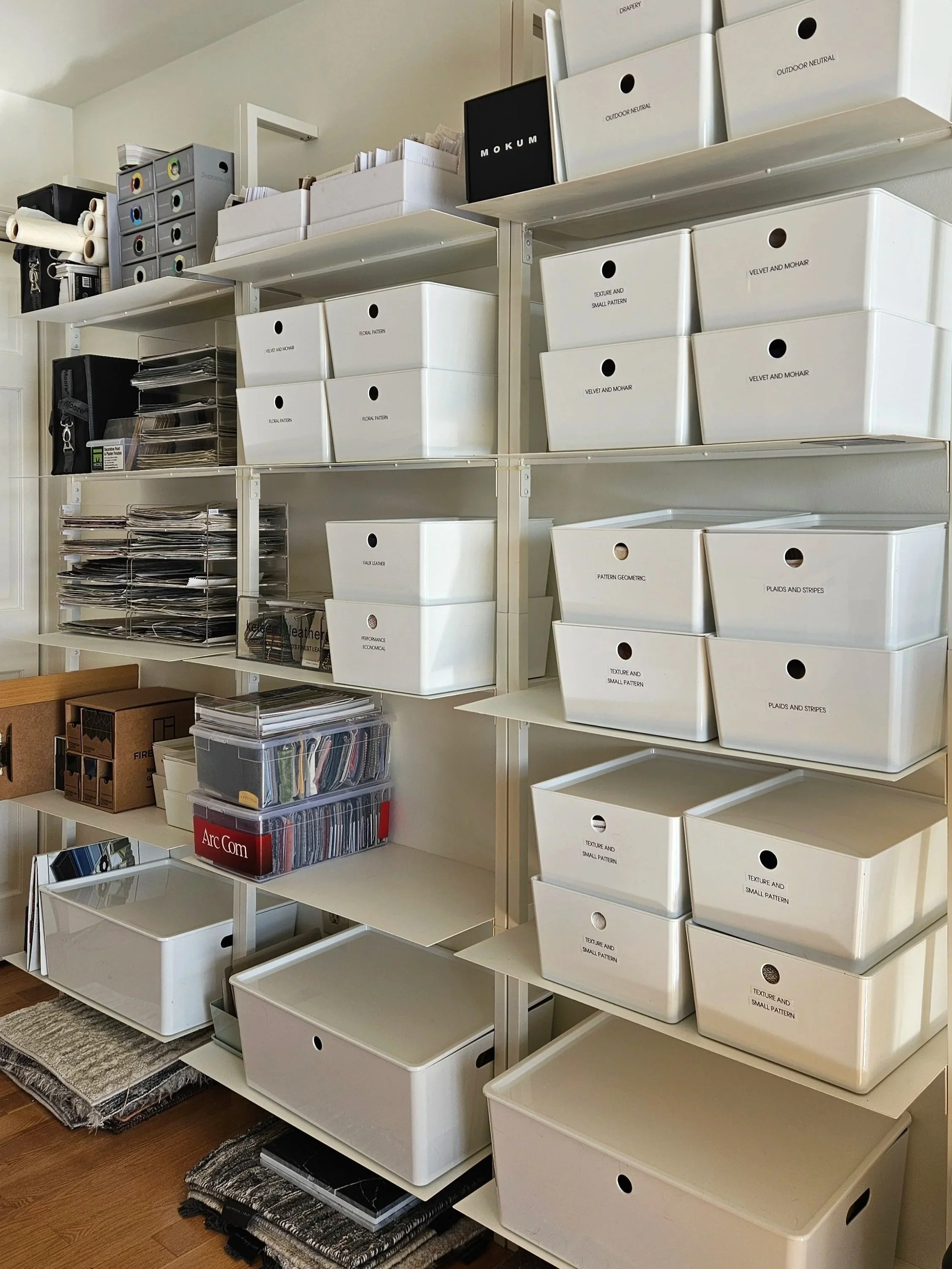 Shelves filled with labeled storage boxes and binders for organizing fabric swatches, patterns, and design materials, located in a workspace or design studio.