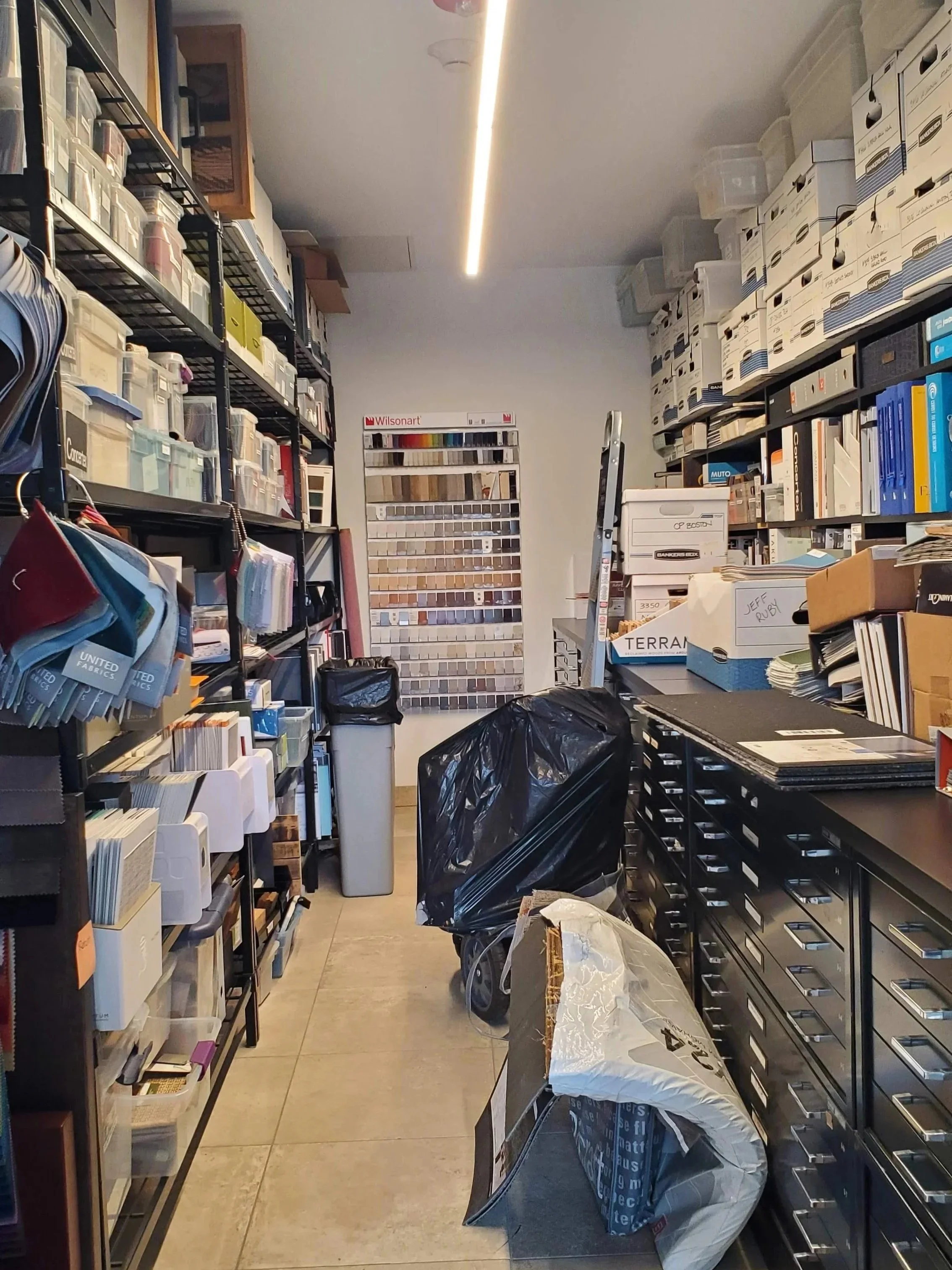 A cluttered storage room with shelves filled with boxes, fabric samples, and files. A ladder and various bags and boxes are on the floor, with a bright overhead light illuminating the space.