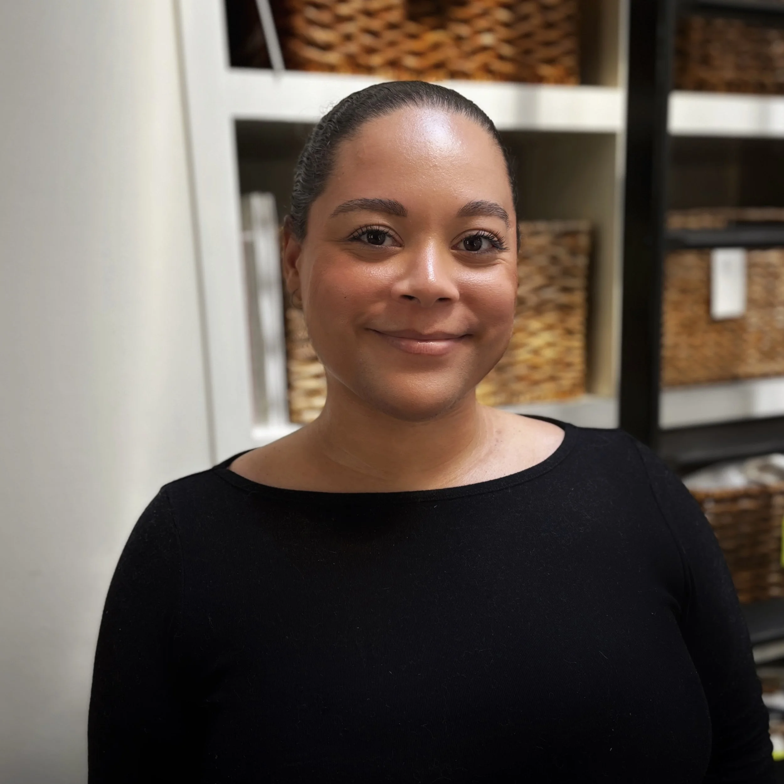 Alexis Dawson, director of operations, smiling, wearing a black top, with woven baskets and shelving in the background.