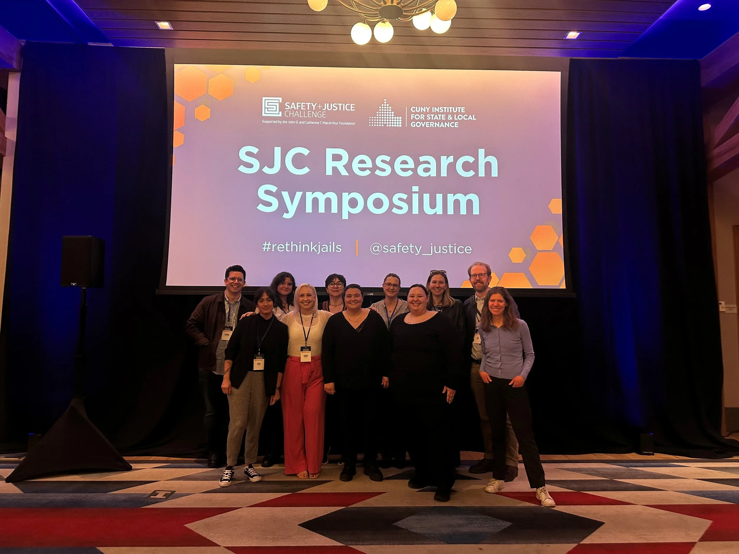 CUNY ISLG staff members smile together in front of a screen that says "SJC Research Symposium"