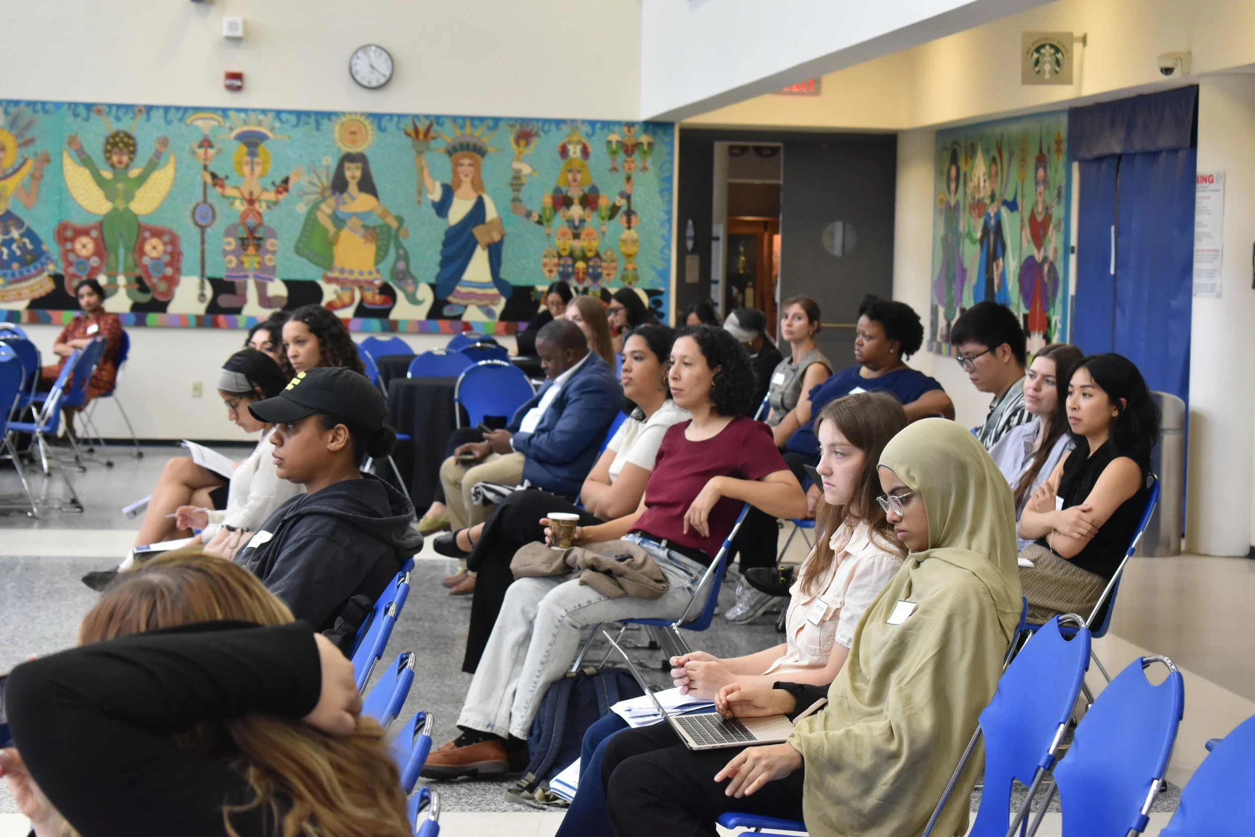 Civic Engagement Fellows, Campaign Finance Board staff, and CUNY ISLG staff sit in blue chairs in a room with colorful murals on the wall, listening to a presenter out of frame.