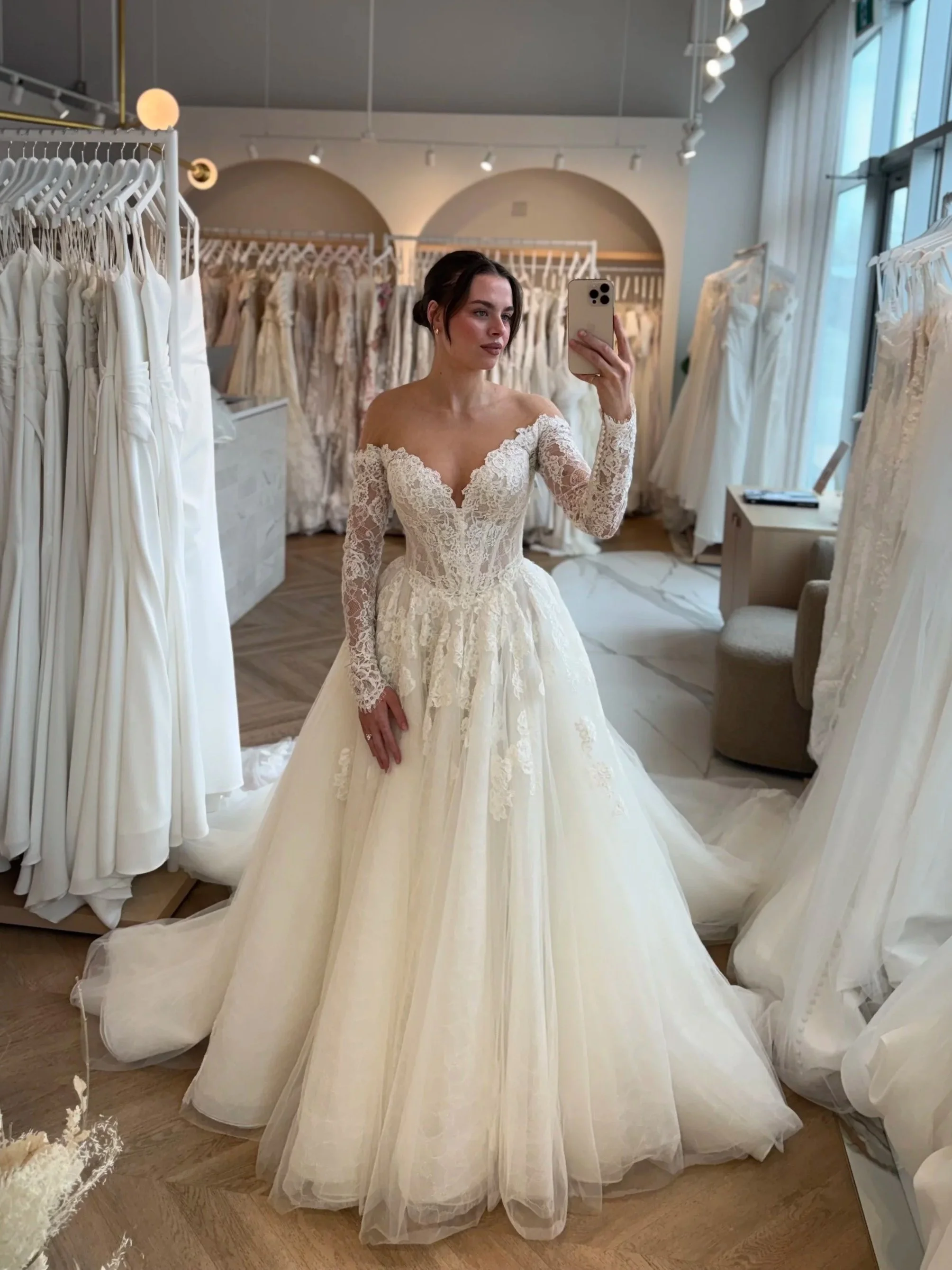 A bride taking a selfie at The Bridal Boutique wearing  Martina Liana 1822. Chantilly lace , Basque waist , off the shoulder long sleeve ballgown. With beautiful dresses in the backgroud