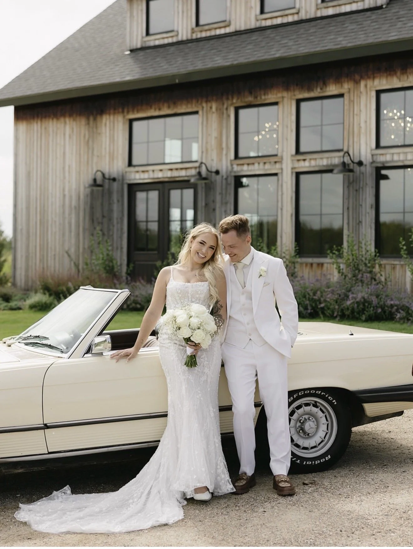 Real bride from The Bridal Boutique holding a bouquet leaning against a vintage car with her husband in an all ivory tux